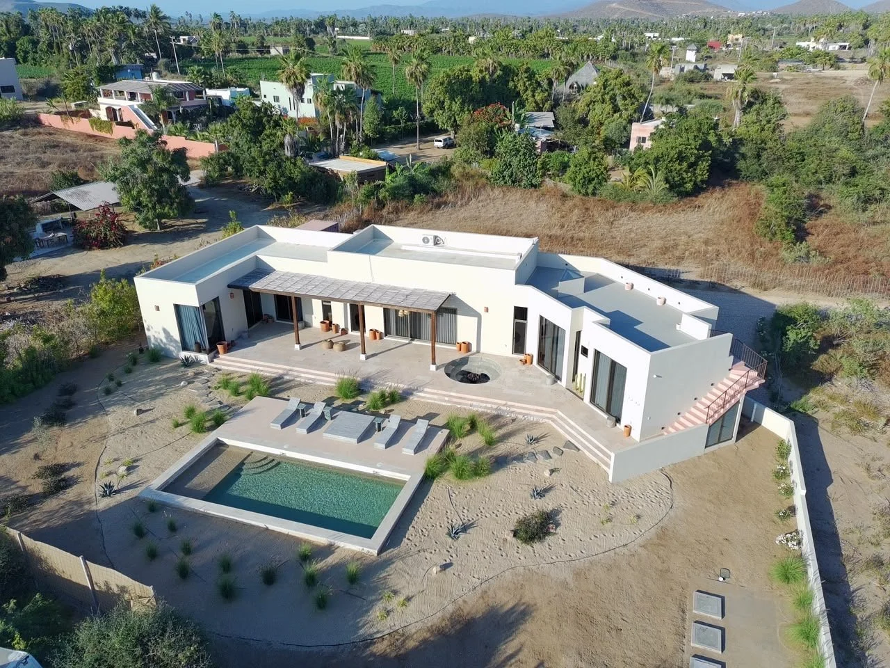 A modern white house with a flat roof, outdoor seating area, rectangular swimming pool, and minimalist landscaping in a semi-dry yard, surrounded by trees and other residential buildings in the background.