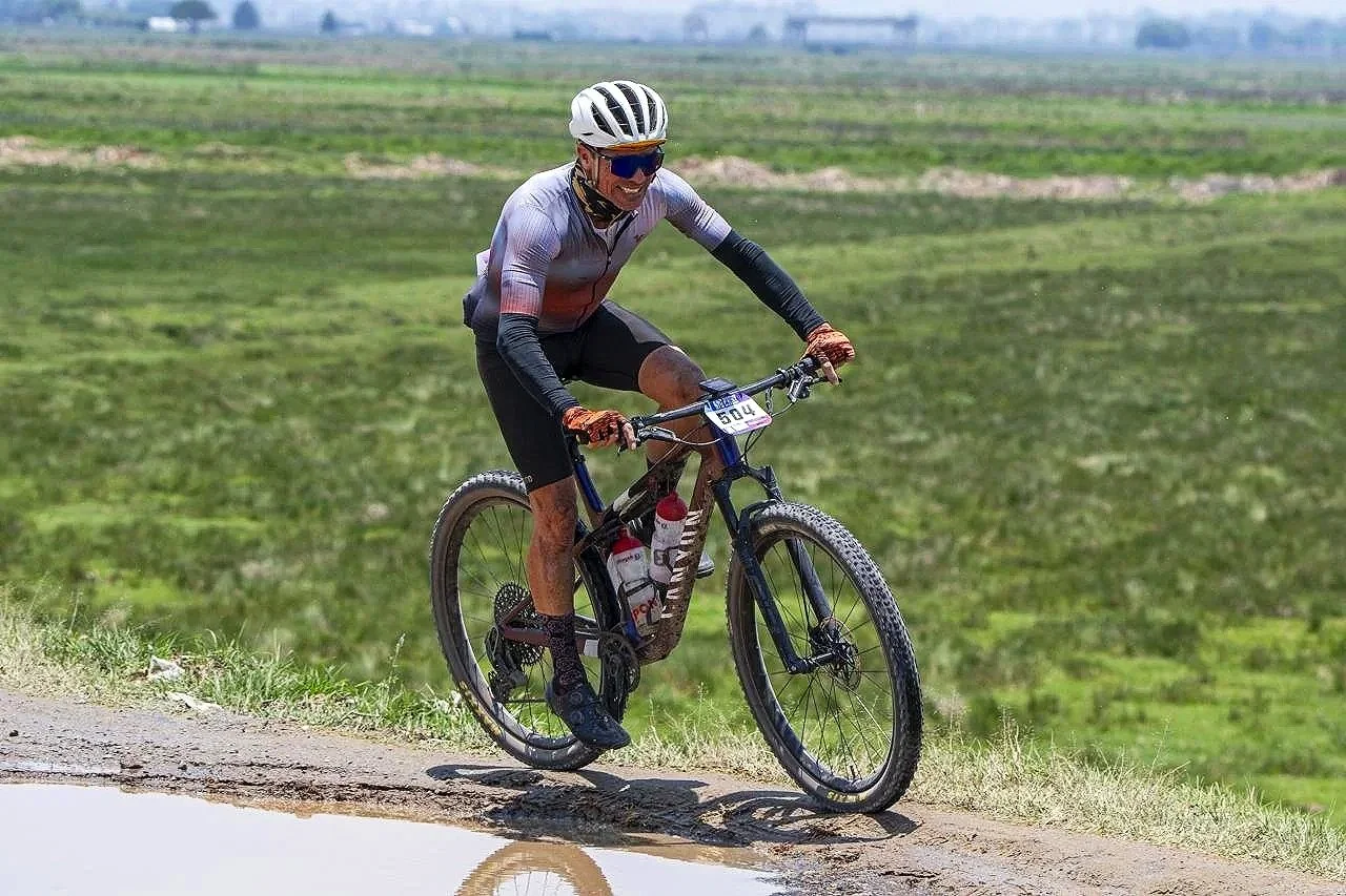 A cyclist wearing a helmet, sunglasses, and sports attire rides a mountain bike on a dirt trail in a green, open landscape.