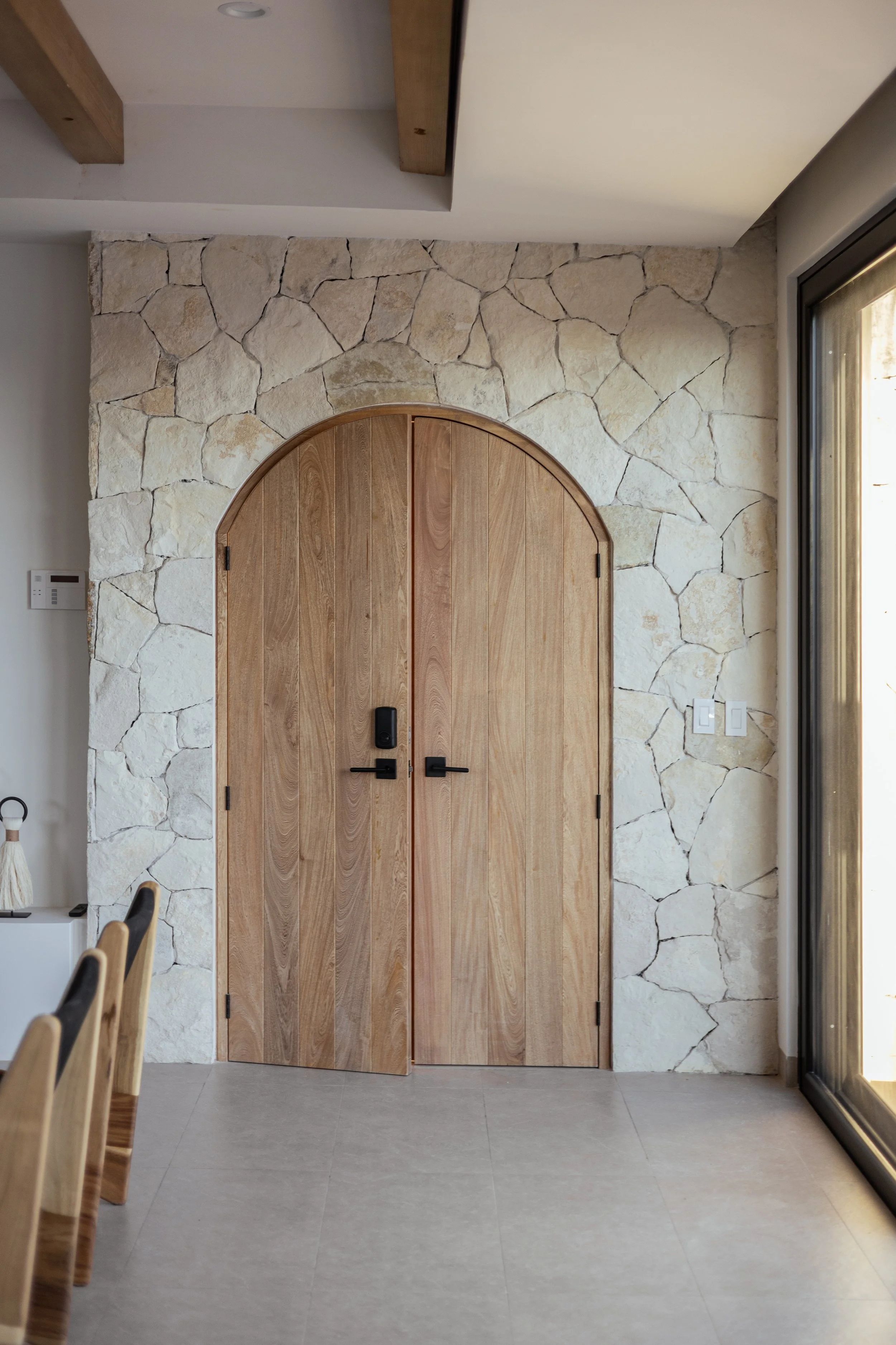 Interior view of a house entrance with a wooden arched door, a stone wall surrounding the door, a sliding glass door to the right, and a tile floor. Part of a wooden dining table and chairs are visible on the left side.