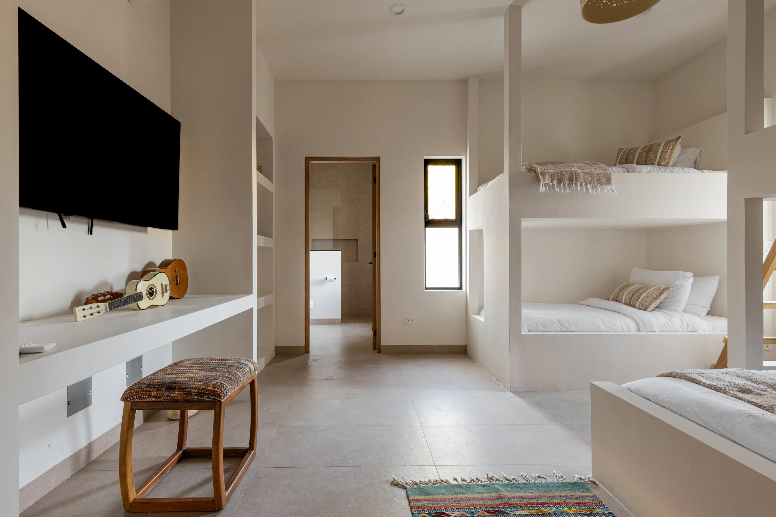 A minimalist bedroom with built-in bunks, a white wall-mounted shelf with two ukuleles, a colorful bench, a small rug, and narrow windows letting in natural light.