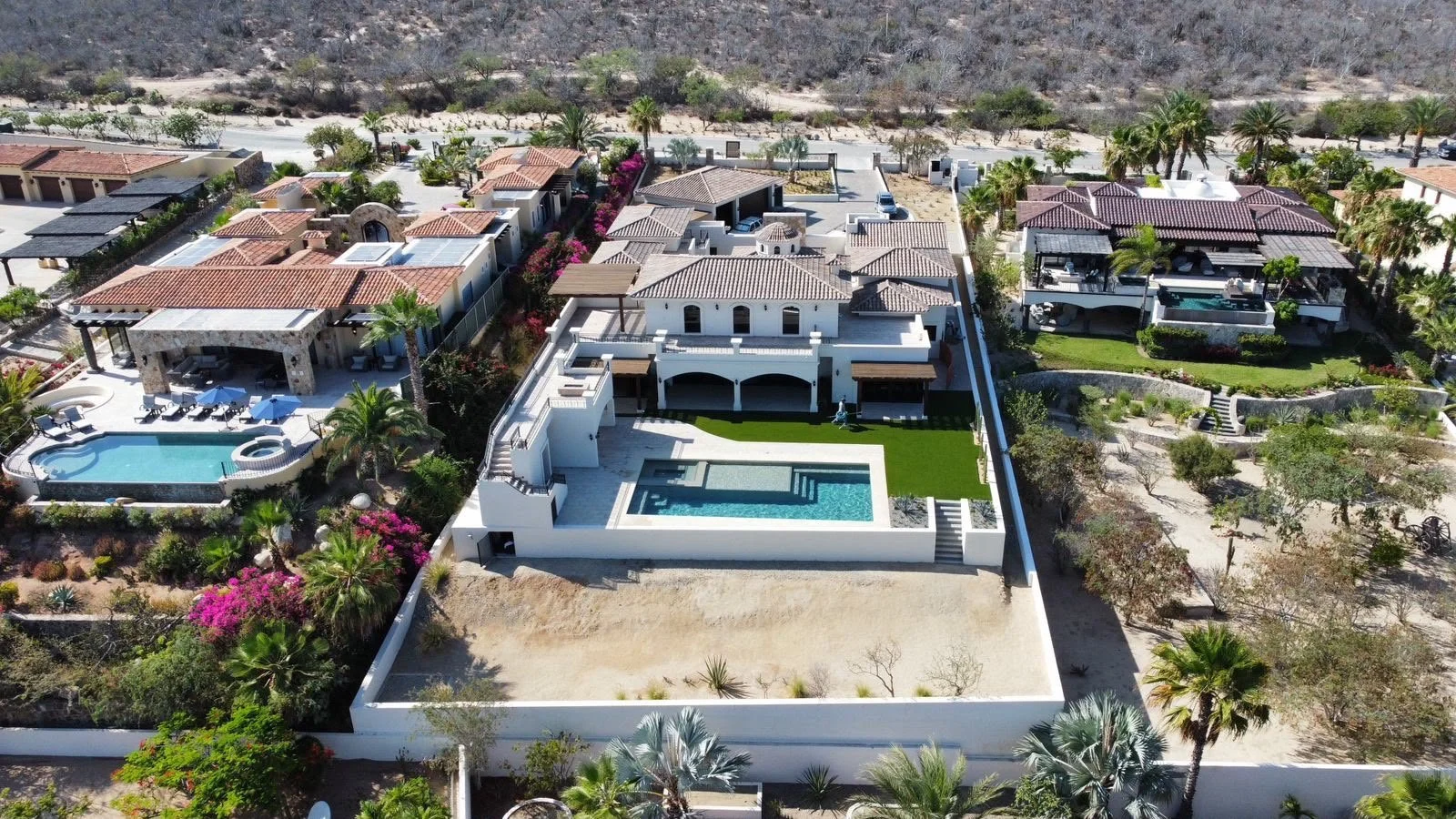 Aerial view of a luxurious neighborhood with multiple houses, including a modern white mansion with a swimming pool in the backyard, surrounded by lush greenery, palm trees, and desert landscape in the background.