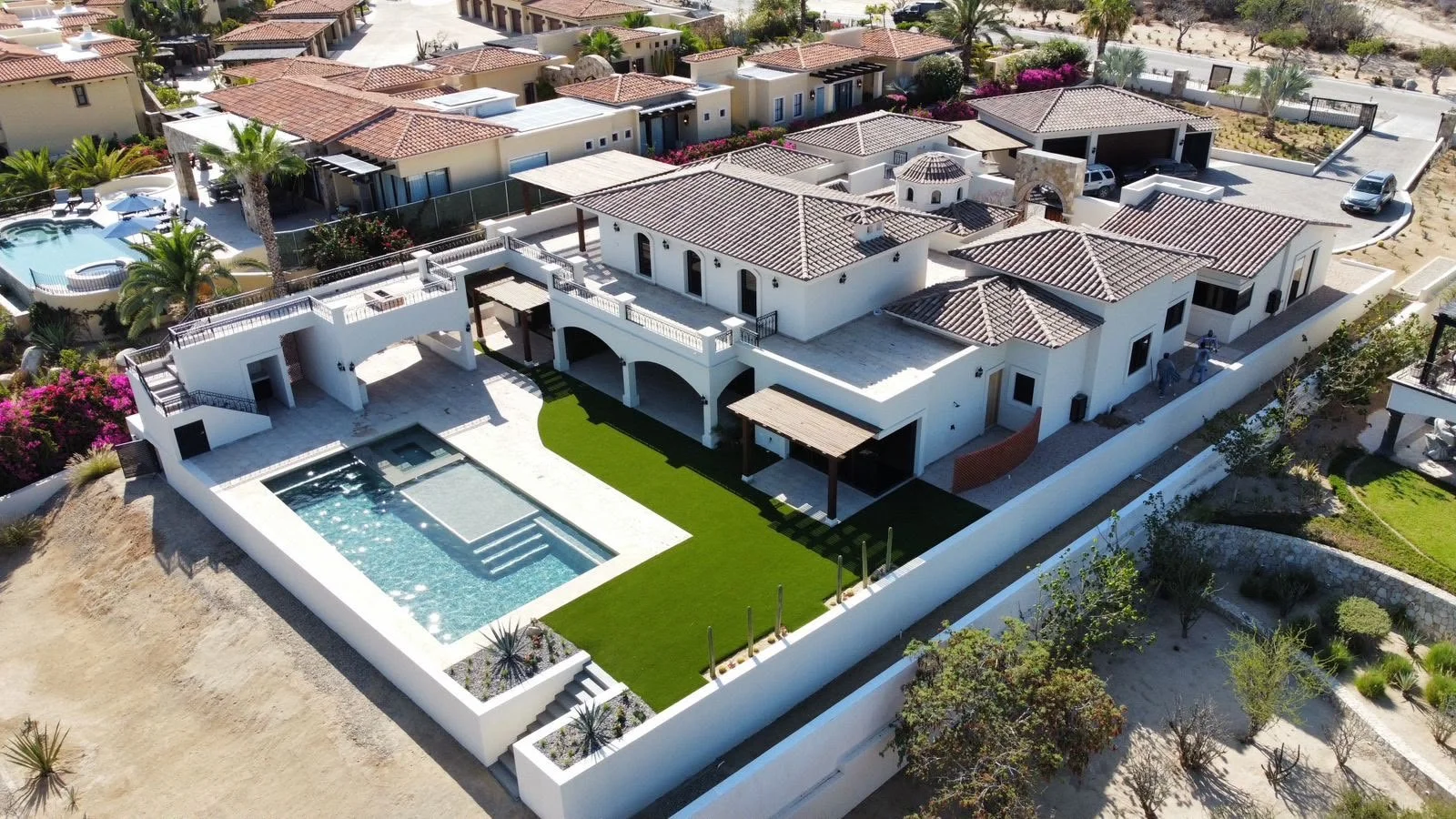 Aerial view of a large, modern white house with a swimming pool and landscaped yard, surrounded by a white boundary wall and neighboring houses in a desert-like area.
