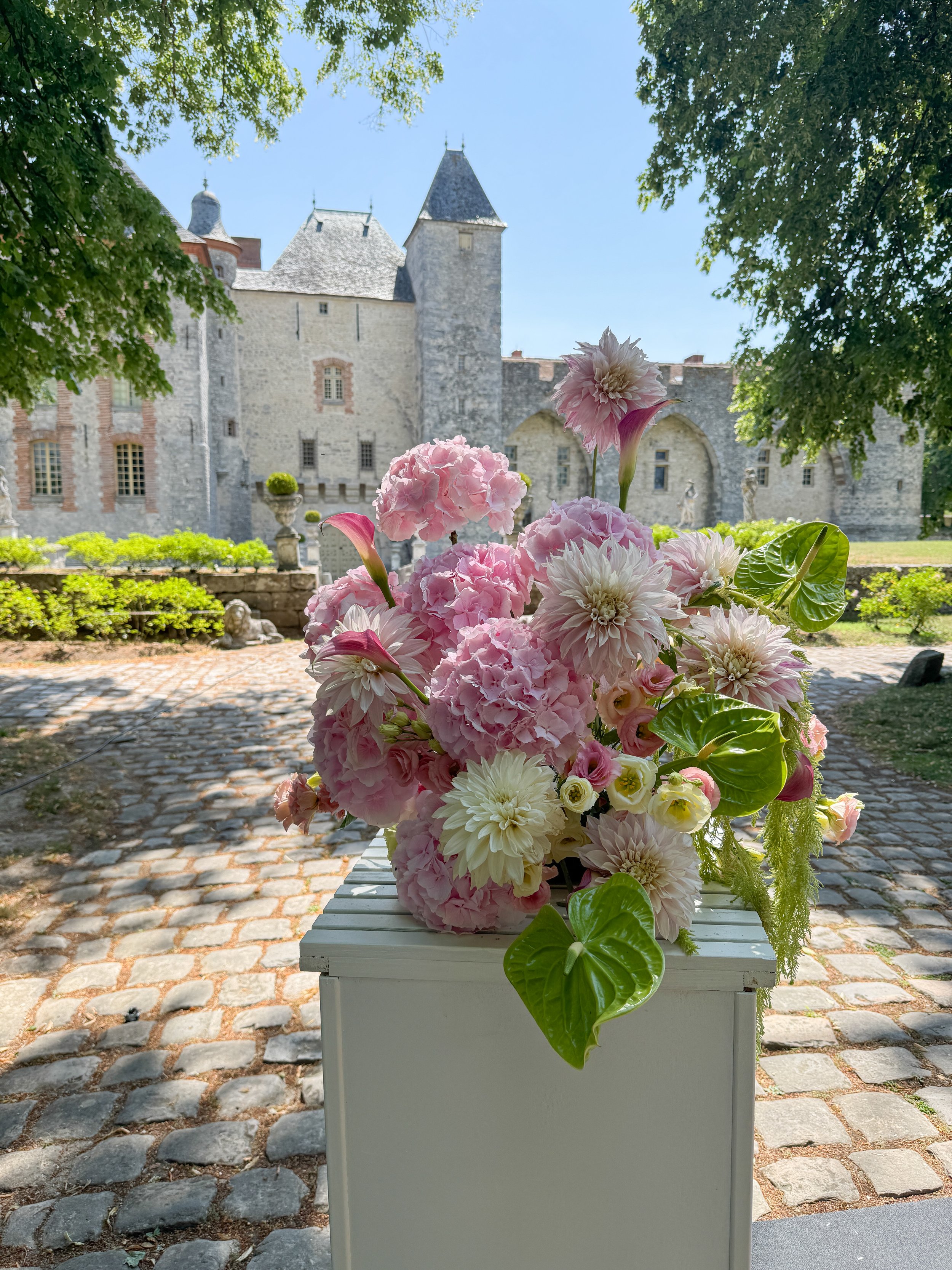 Un arrangement floral de grandes fleurs roses, blanches et violettes est placé sur une table blanche à l'extérieur, avec un château en pierre en arrière-plan et des arbres ombrageant la scène par une journée ensoleillée.