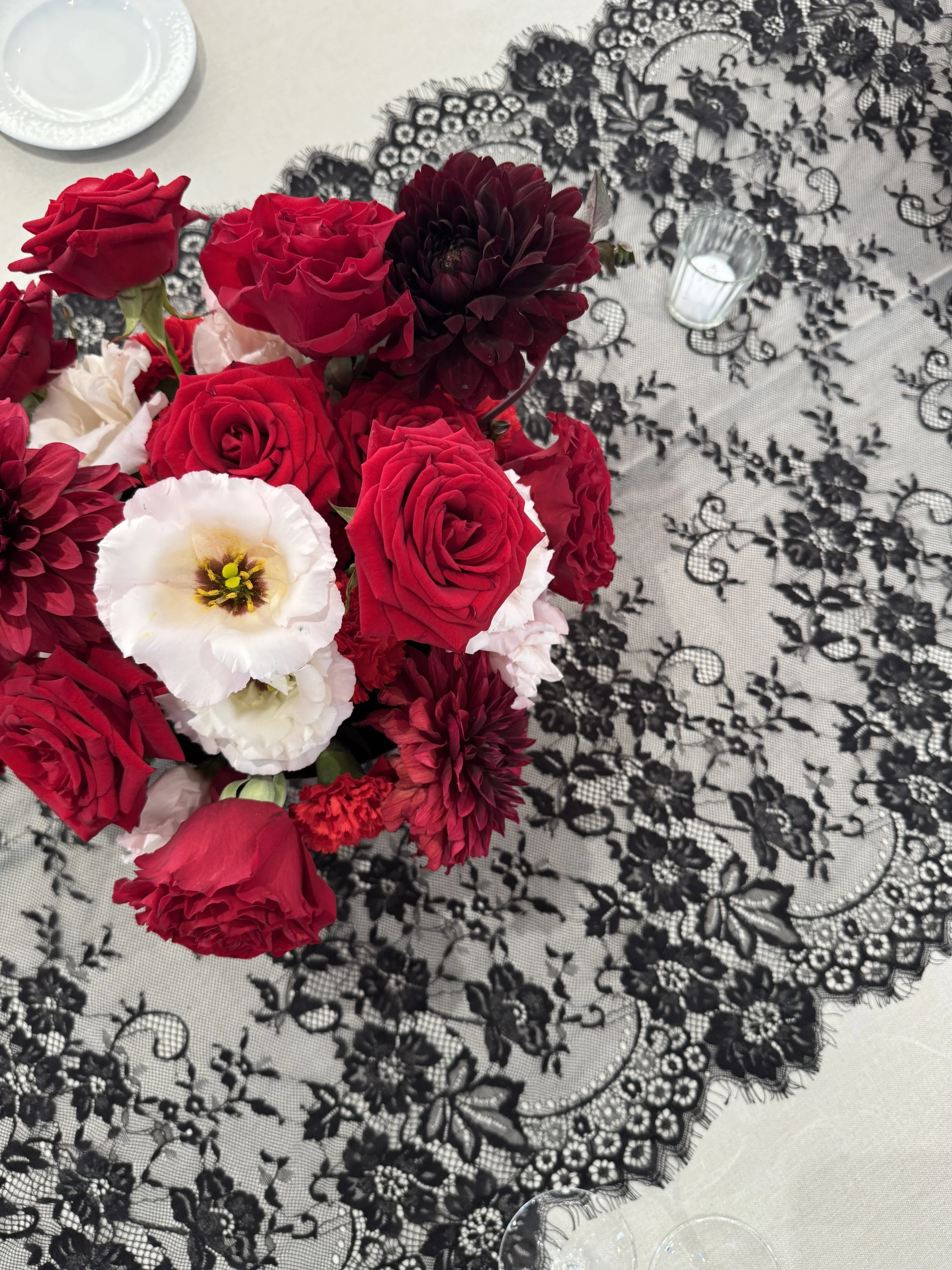 Un bouquet de roses rouges, roses blanches, et d'autres fleurs foncées, placé sur une nappe en dentelle noire avec un motif floral, sur une table avec des assiettes et un bougeoir contenant une bougie