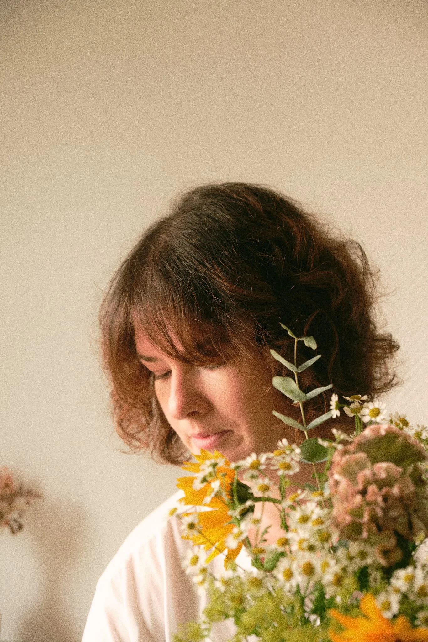 Une femme avec des cheveux courts et bouclés, portant une robe rose clair, tenant un bouquet de fleurs colorées, face à un mur beige.