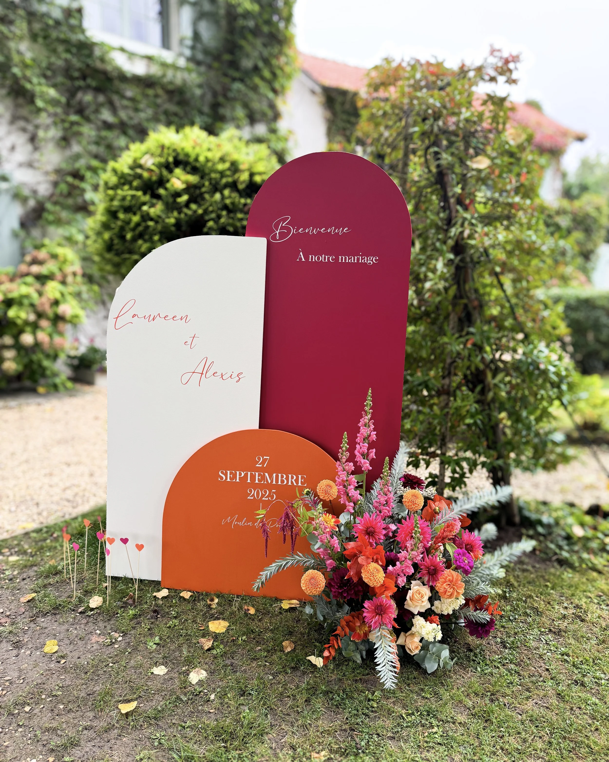 Message de bienvenue pour un mariage, avec un grand bouquet de fleurs colorées en premier plan, textes en français sur des panneaux de couleur rose, blanc et orange, fond de jardin avec végétation et maison en arrière-plan.