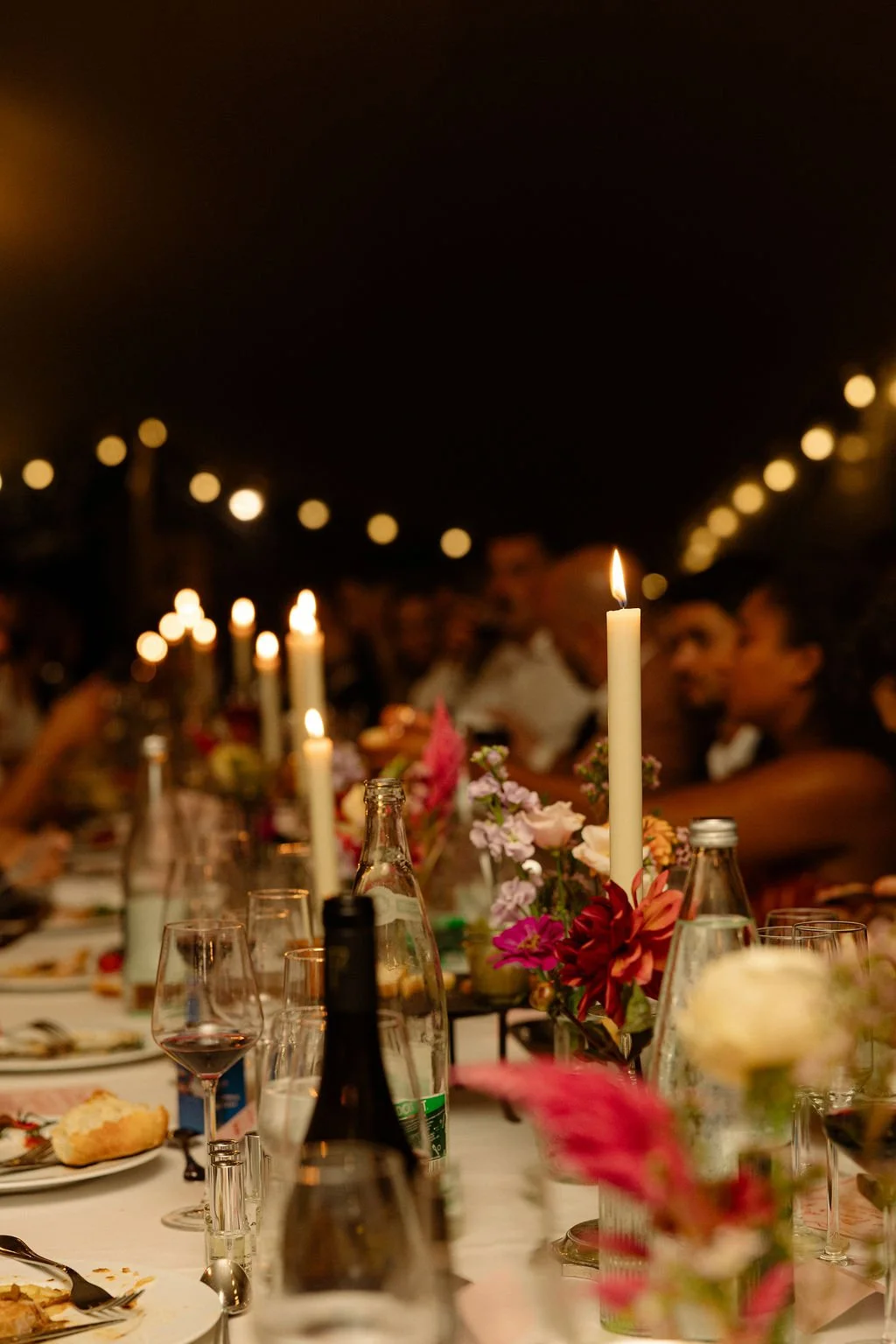 Un dîner en soirée avec un long repas, lampe à chandelle, fleurs colorées, et plusieurs convives. Décor chaleureux et intime avec des lumières suspendues.
