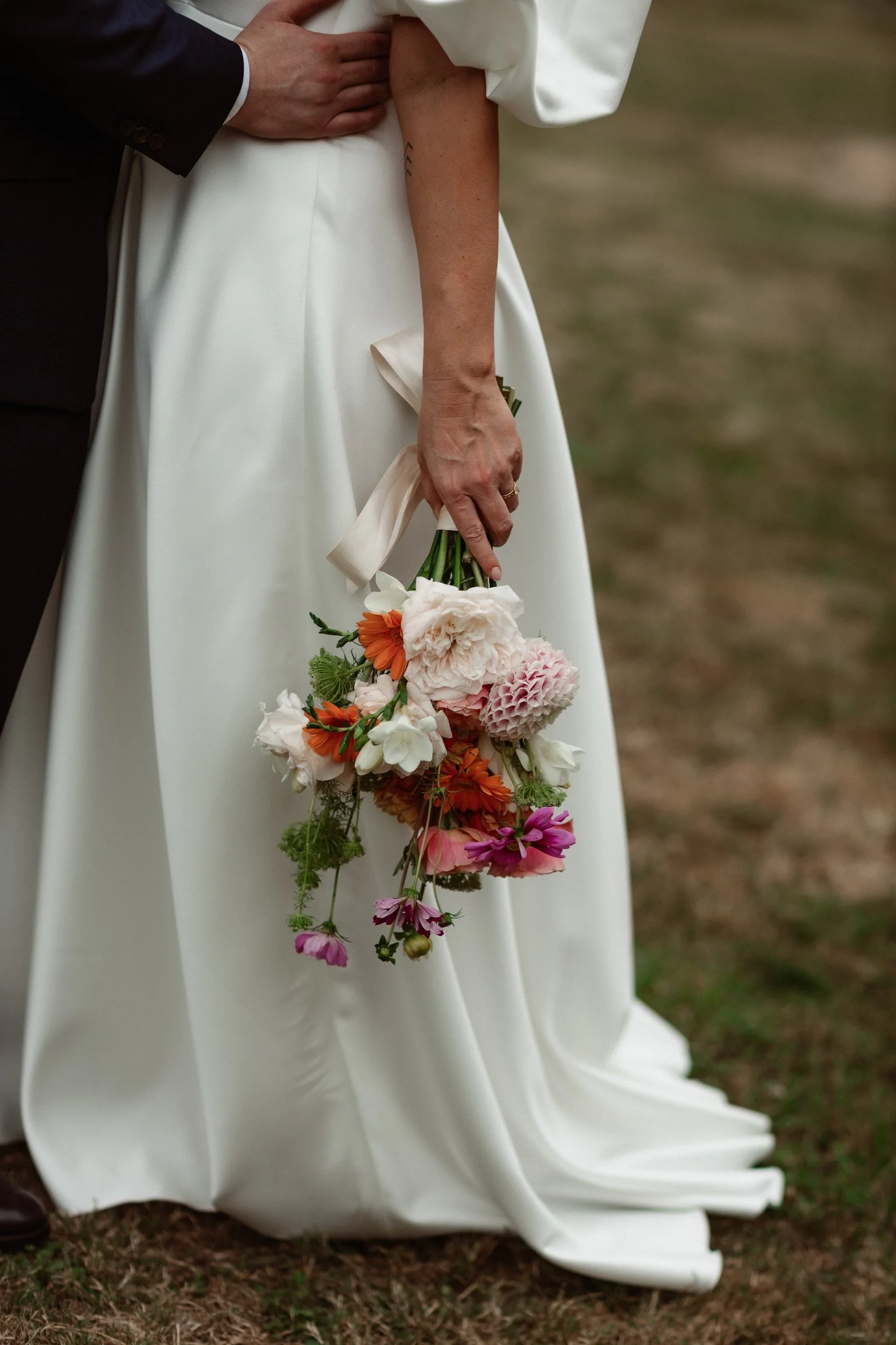 Une femme en robe de mariage blanche tenant un bouquet de fleurs colorées, avec un homme en costume partiellement visible, dans un cadre naturel.