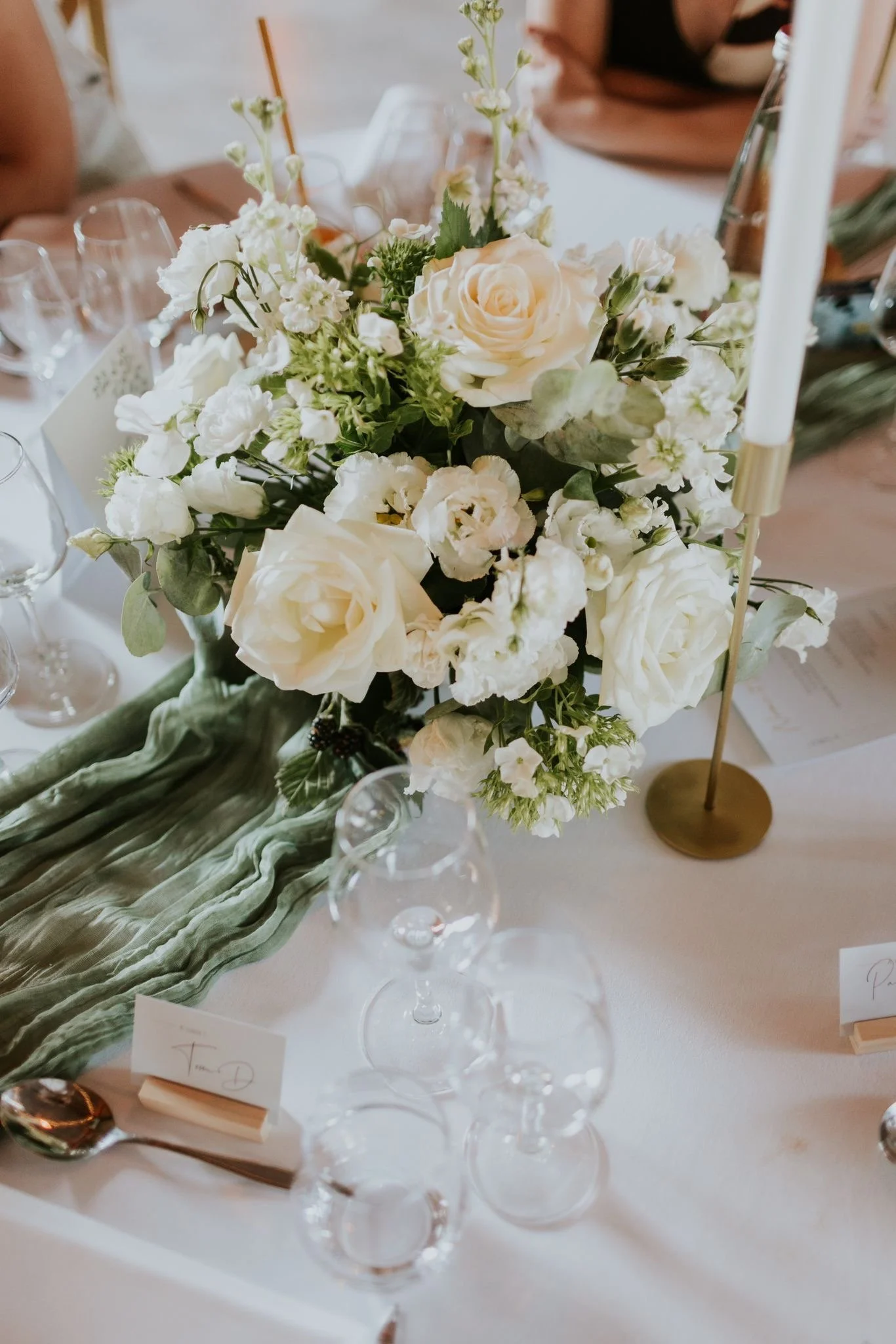 Un arrangement floral composé de roses blanches et d'autres fleurs blanches, placé sur une table avec des verres à vin en cristal et une nappe blanche, ambiance d'événement formel ou de mariage.