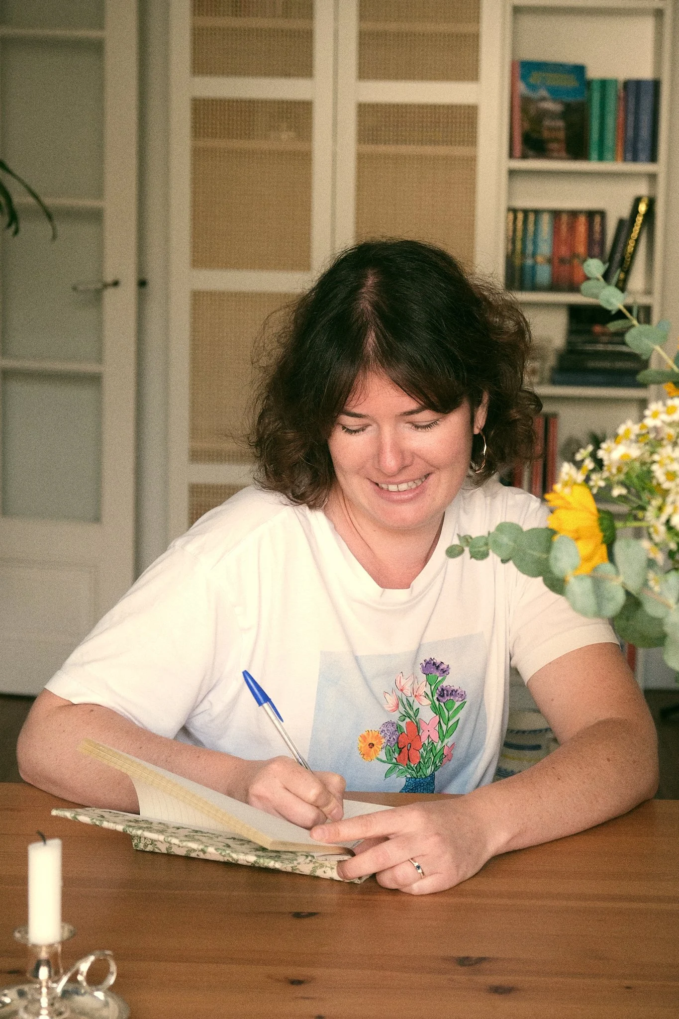 Une femme souriante écrit dans un carnet, assise à une table en bois avec un bouquet de fleurs en face d'elle.