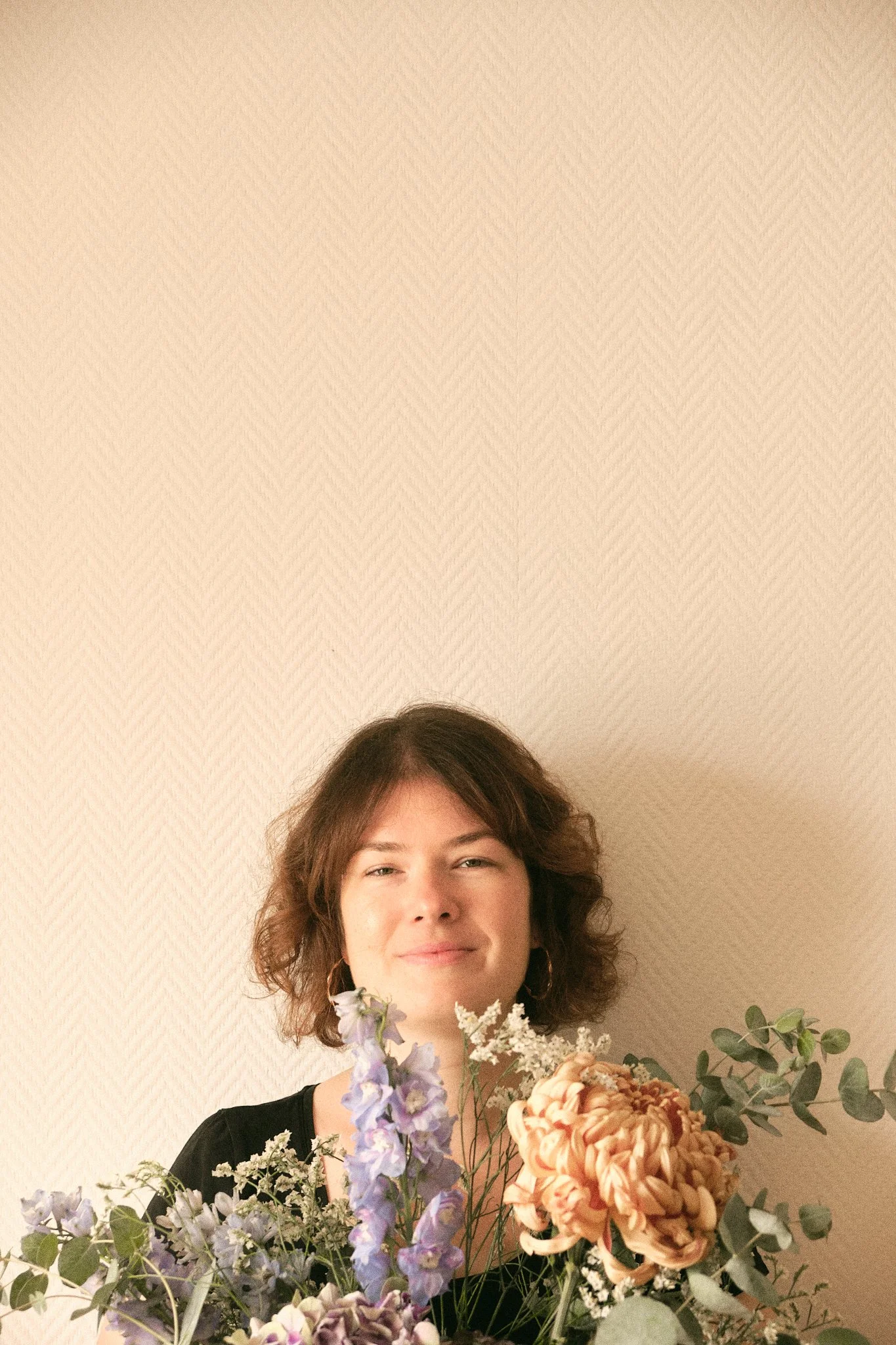 Une femme avec des cheveux bouclés, portant un haut noir, se tenant devant un mur beige, entourée de plusieurs bouquets de fleurs aux teintes pastel.
