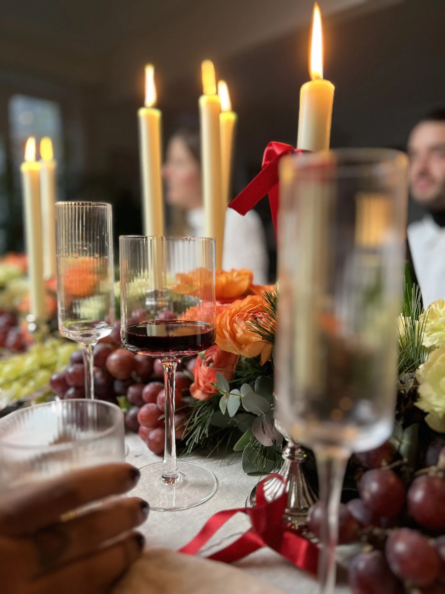 Une table de fête décorée avec des bougies allumées, des verres de vin rouge, des fleurs orange et des grappes de raisin, avec deux personnes floues en arrière-plan.