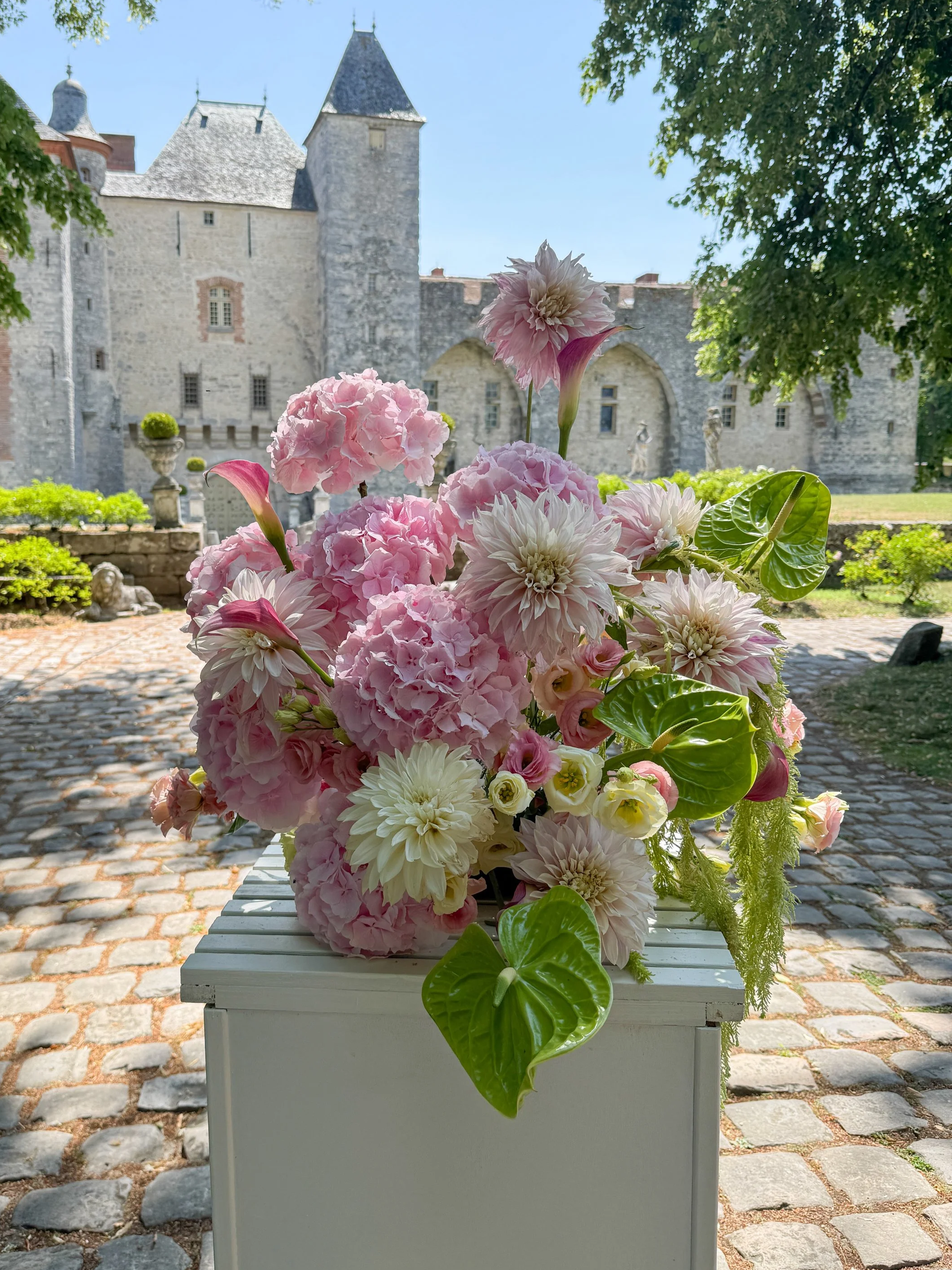Un arrangement floral de fleurs roses, blanches et vertes placé sur une colonne blanche dans une cour pavée devant un château médiéval en pierre avec des tours et des murs.