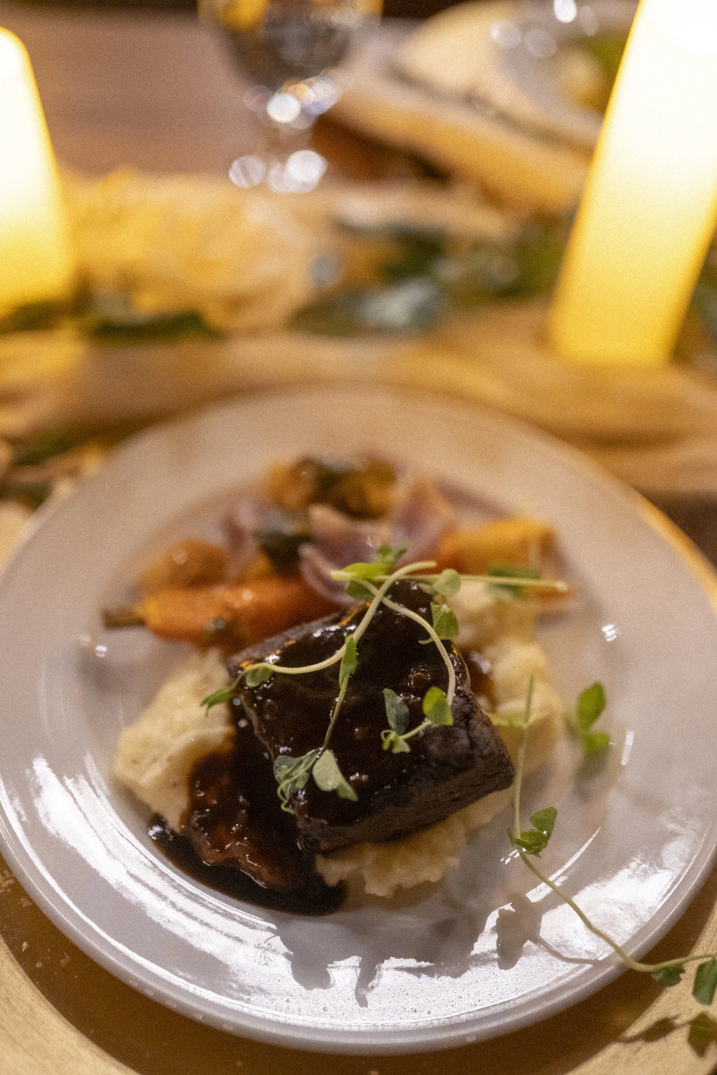 A plated gourmet meal featuring a piece of meat with sauce, served over mashed potatoes with vegetables in the background, set on a table with a lit candle.