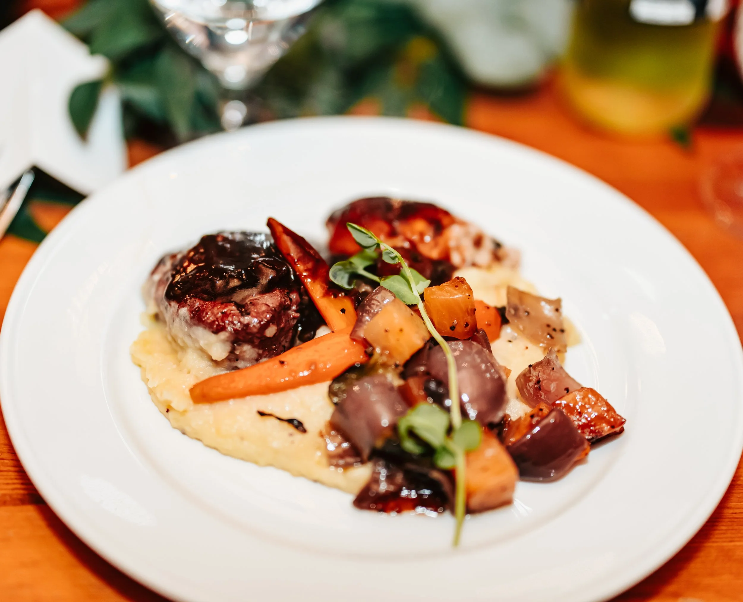 A plated main dish featuring beef with sauce, roasted vegetables, and mashed potatoes garnished with microgreens on a white plate.