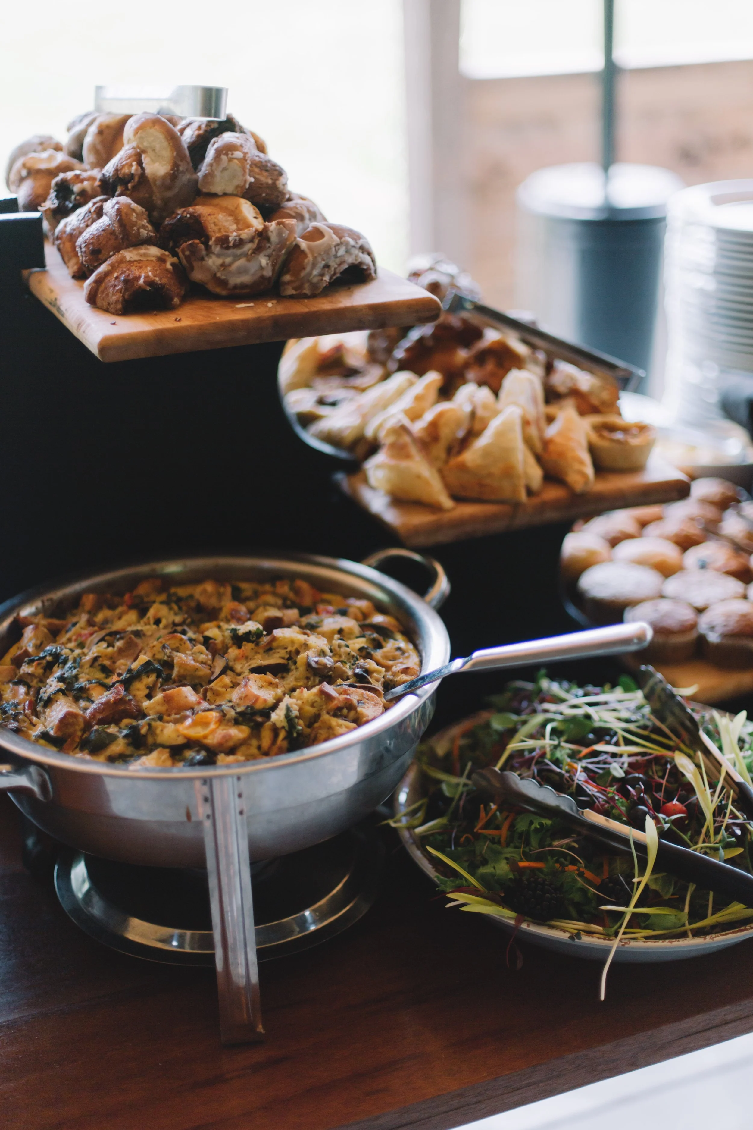 Buffet table with assorted food items including pastries, a vegetable casserole, a mixed green salad, and a plate of cookies.