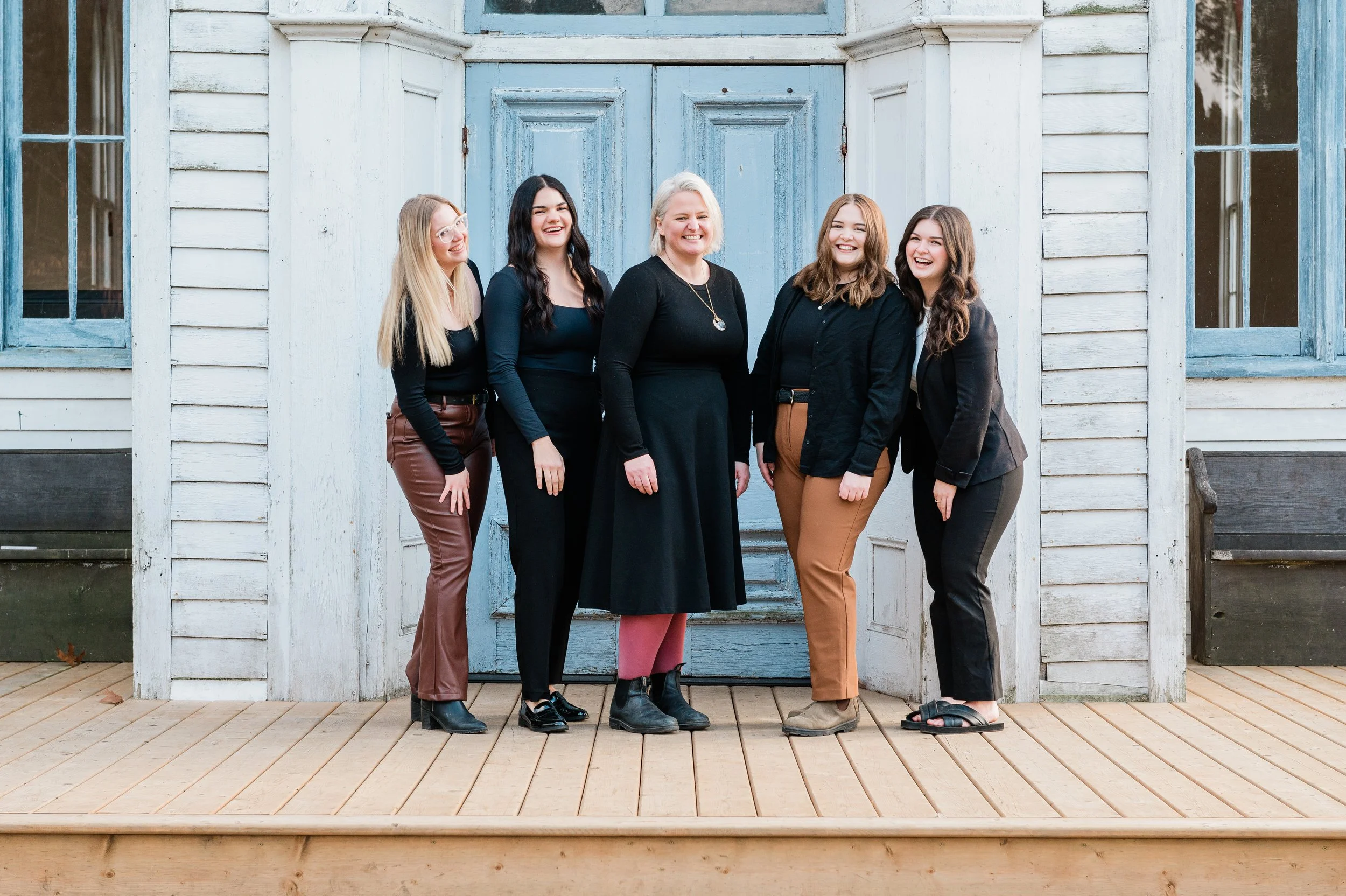 A group of five women standing side by side on a wooden deck, smiling, in front of a rustic blue double door with windows, wearing casual and semi-formal clothing.