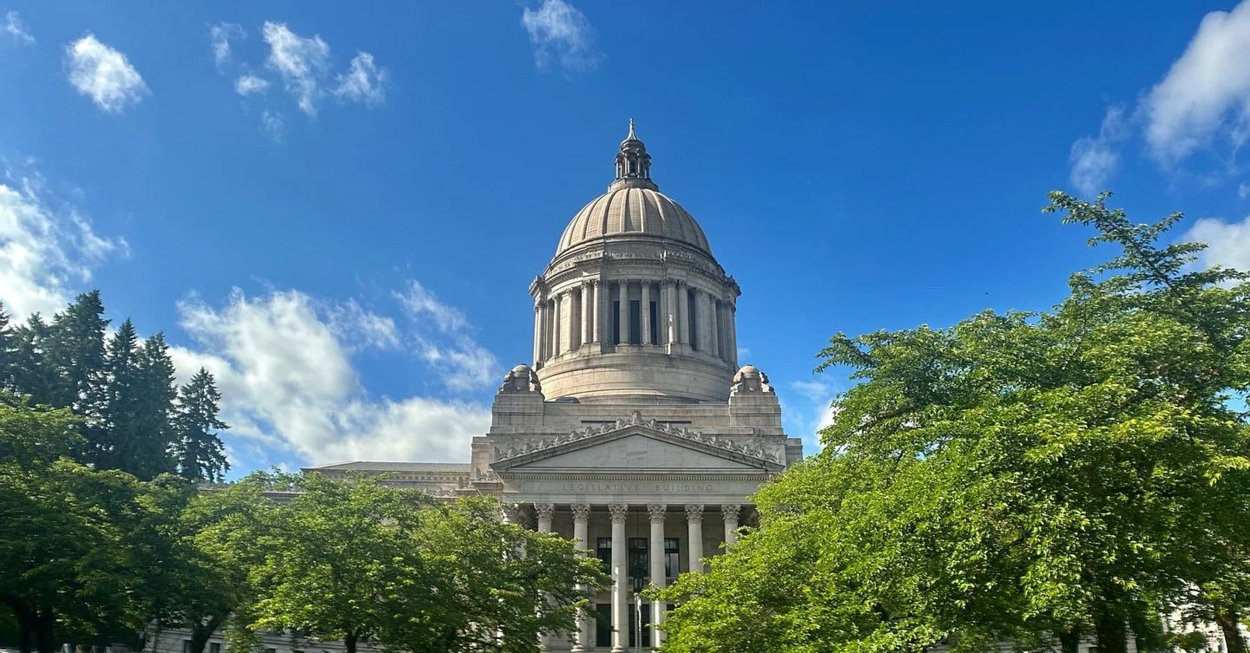 The Capitol building in Olympia, Washington, with a prominent dome, surrounded by green trees and a bright blue sky with some white clouds.