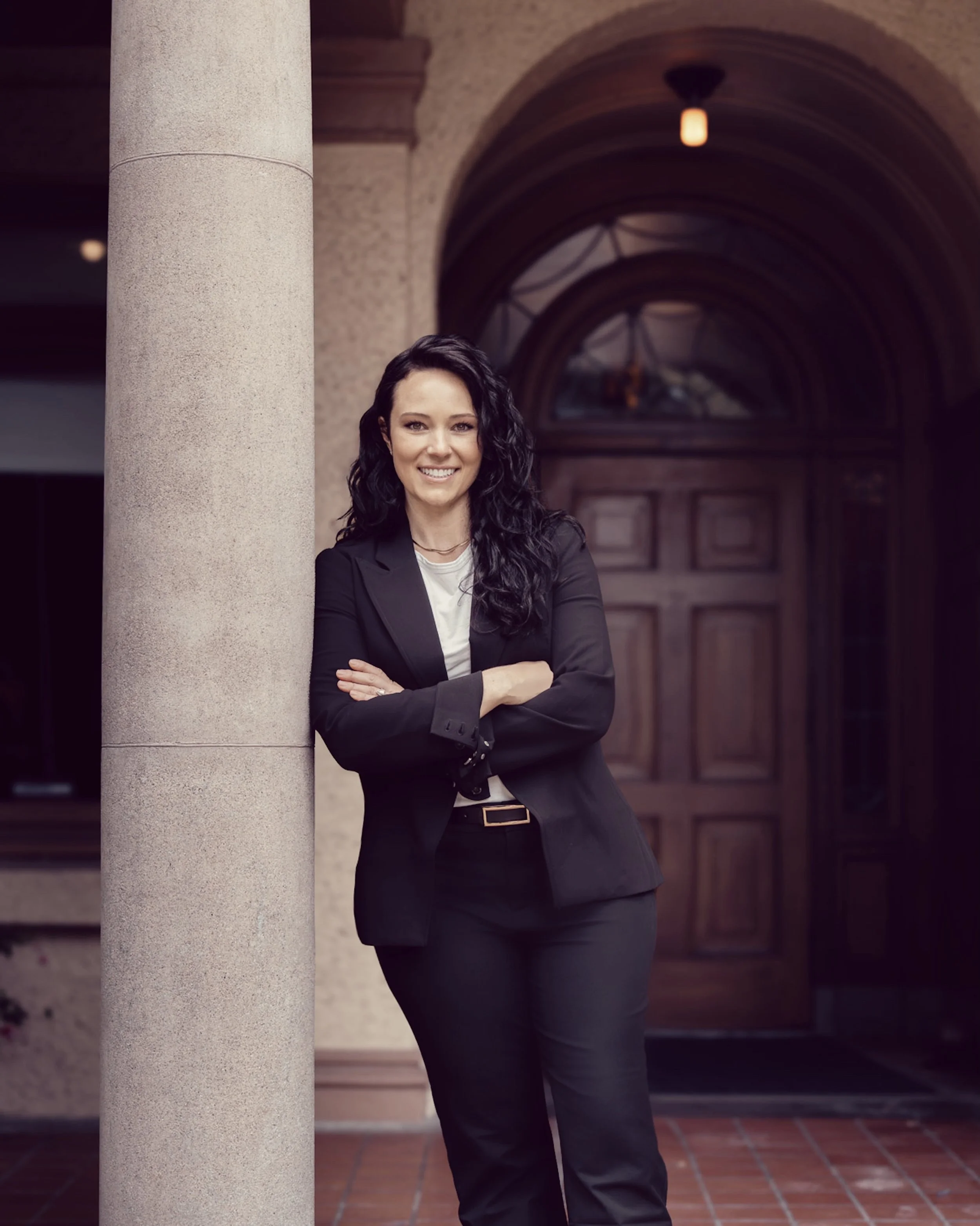 A woman with long curly black hair, smiling, standing outdoors near a beige stone column and a large arched window with wooden doors, dressed in a black blazer and pants with a white top underneath.
