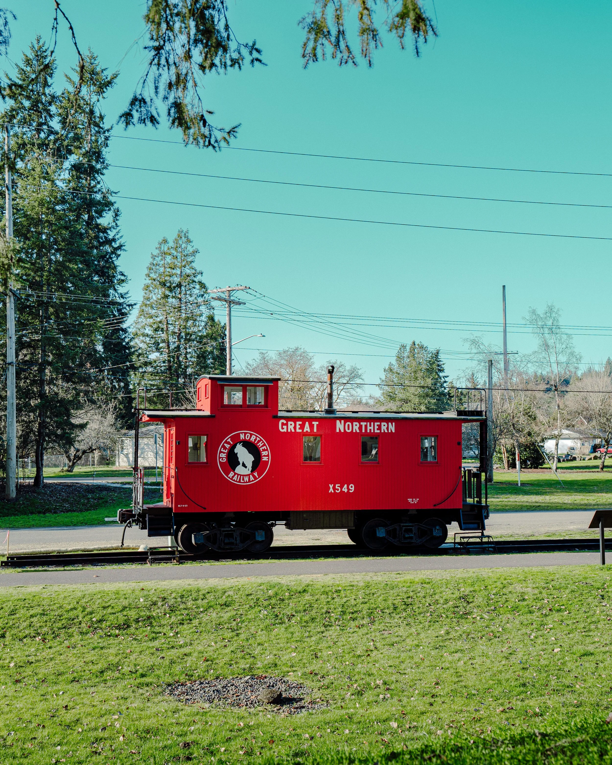 A Tenino red train car with the words "Great Northern Railway" on it, parked on a track outdoors with green grass in the foreground. There are trees and utility poles in the background and overhead wires for the train. The sky is clear and blue.