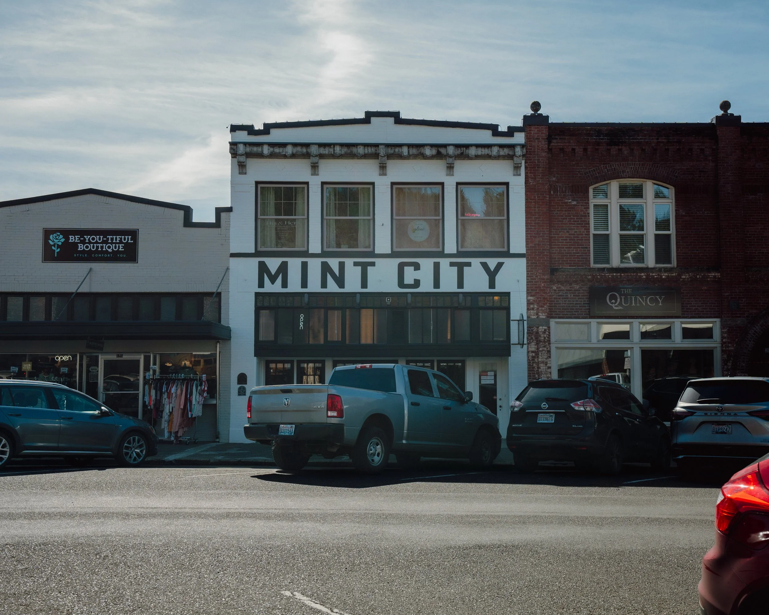 Street view of downtown Chehalis storefronts with cars parked in front, including a boutique named 'BE-YOU-TIFUL BOUTIQUE' and other shops such as Quincy with visible signage, under a partly cloudy sky.