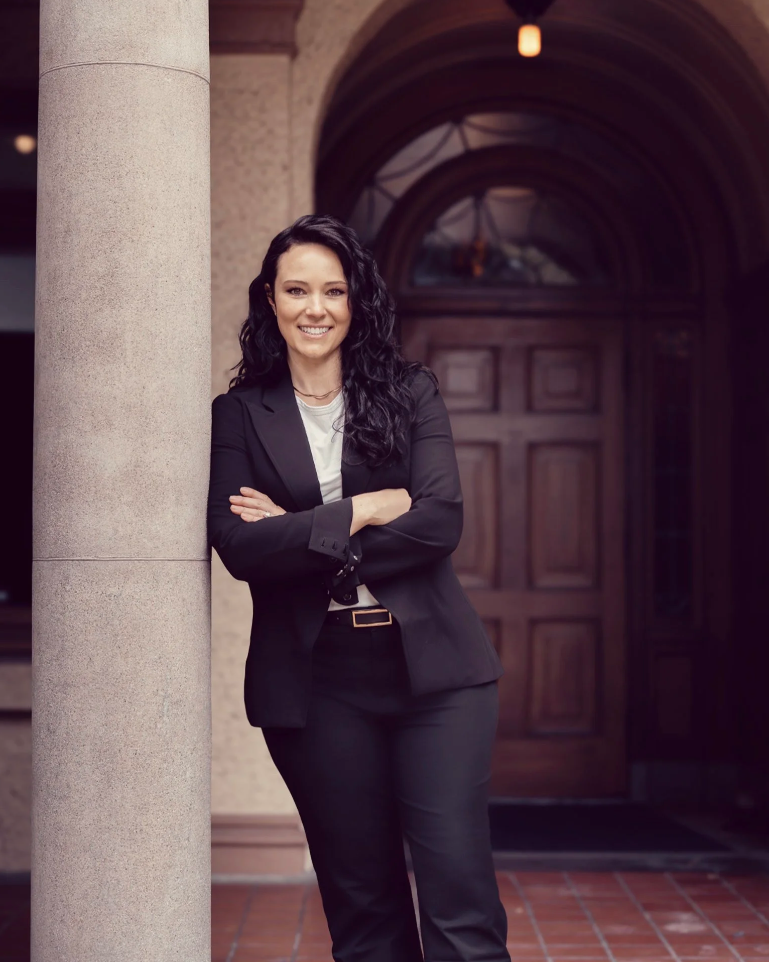 A woman in a black suit smiling and standing outdoors near a light-colored pillar with a wooden door and arched windows in the background.