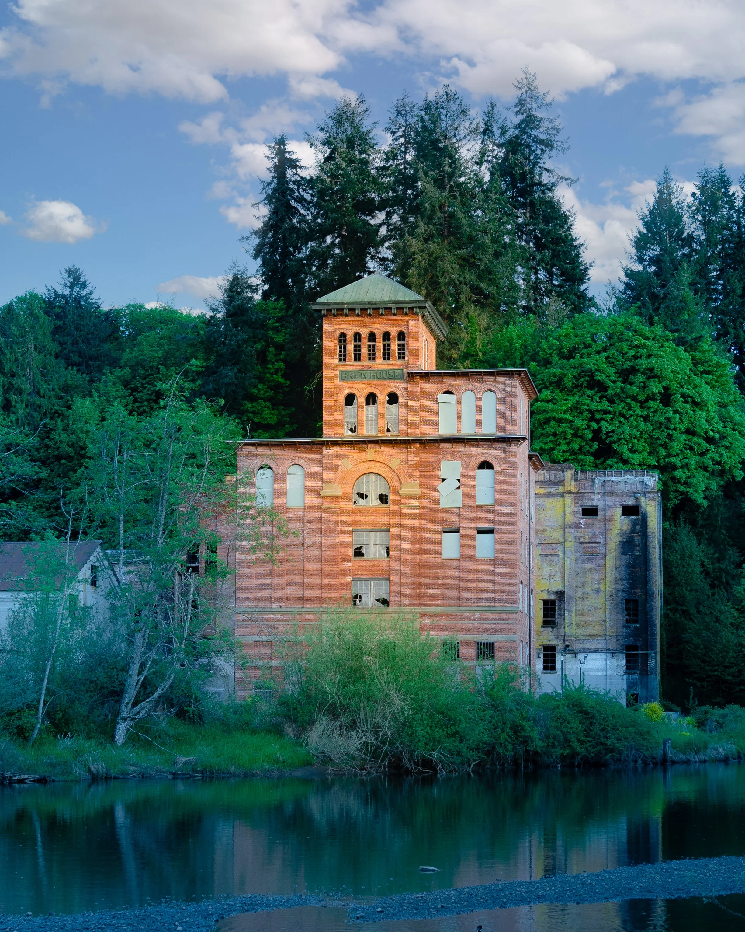 Old brick building of the historic Tumwater Olympia Brewery Company labeled 'Brew House' with broken windows, surrounded by trees and next to a river under a partly cloudy sky.