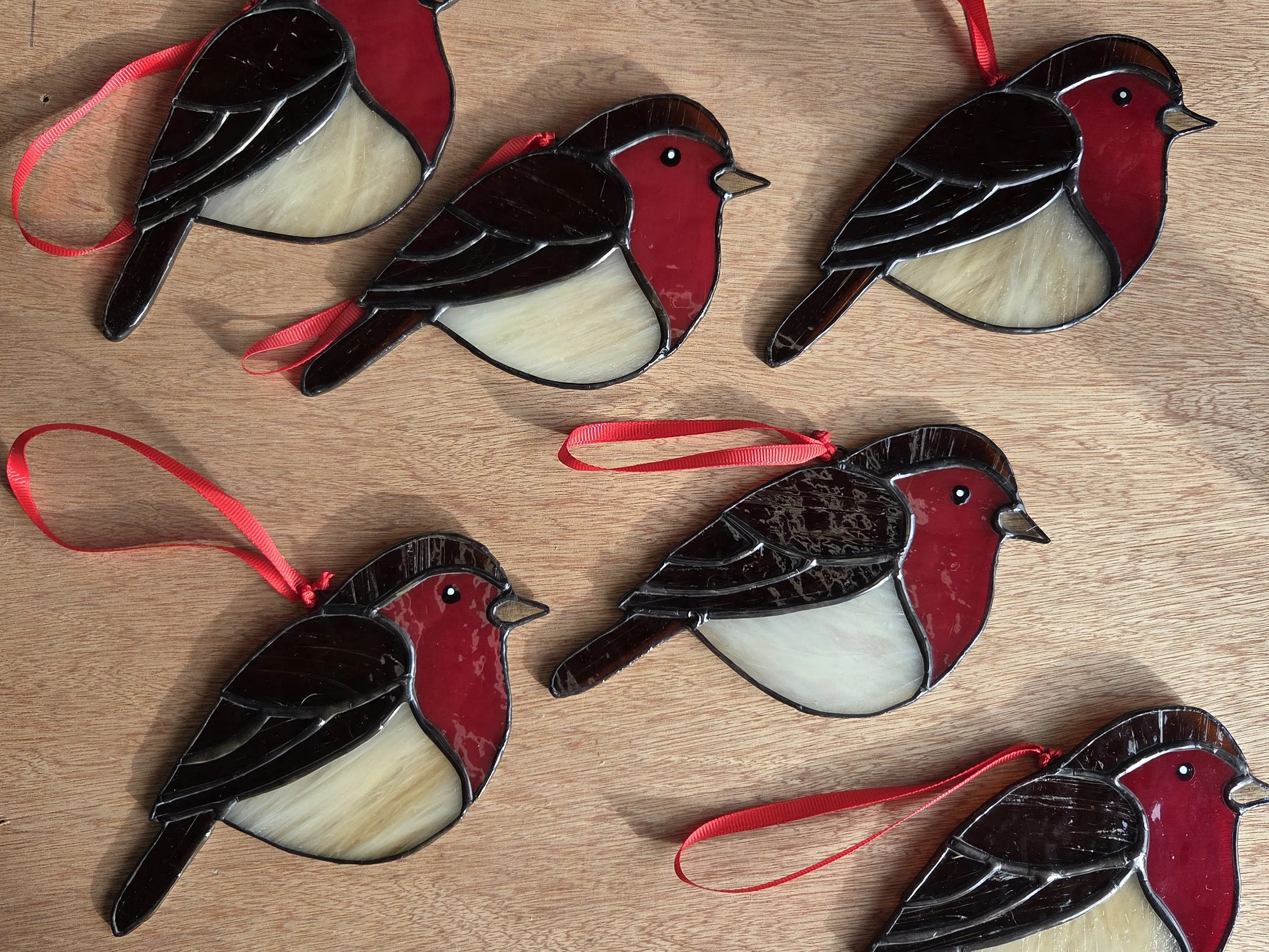 Several robin stained glass suncatchers with red breasts and red hanging ribbons, arranged on wooden background.