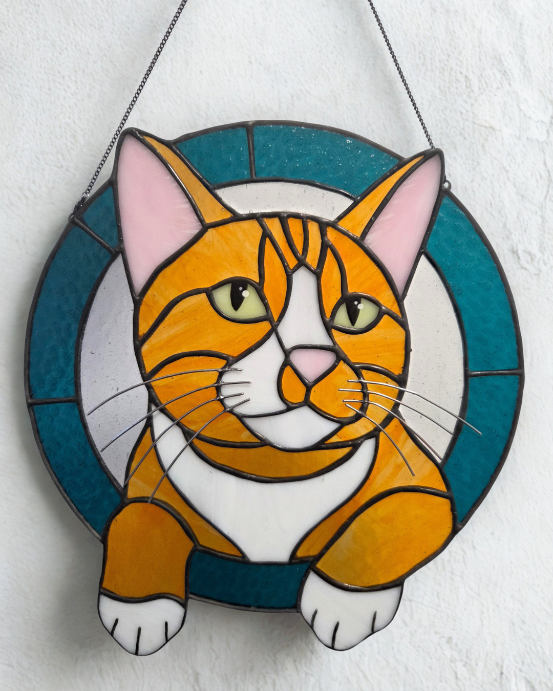 Colorful stained glass art depicting a sitting orange tabby cat with green eyes and pink ears, framed by a circular teal and gray background, hung on a white wall.