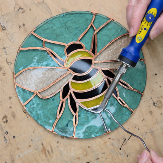A stained glass circular art piece in progress, featuring a bee design with colored glass pieces and copper foil, on a wooden work surface, with a hand using a soldering iron.