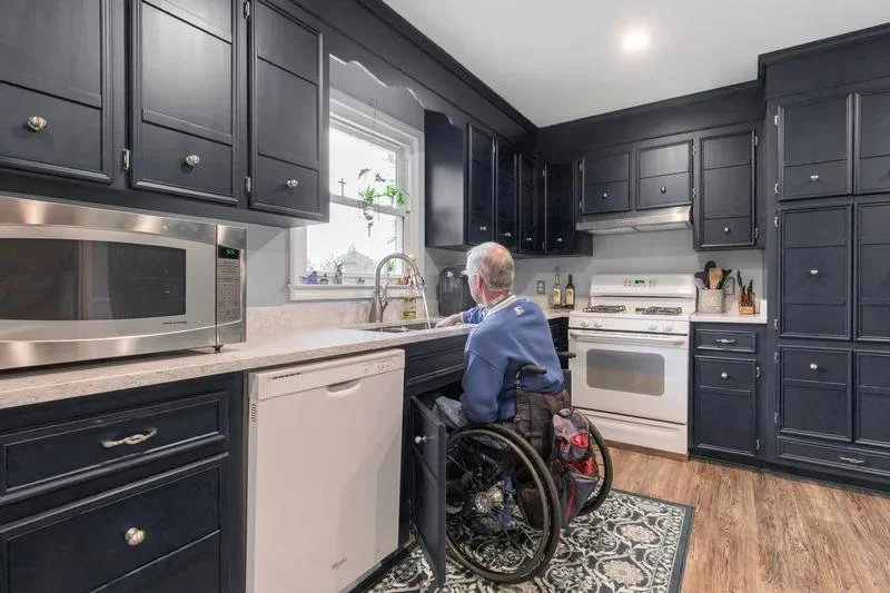 Elderly man in a wheelchair preparing food at a kitchen sink with dark blue cabinets, a microwave, a white stove, and a dishwasher in the kitchen.