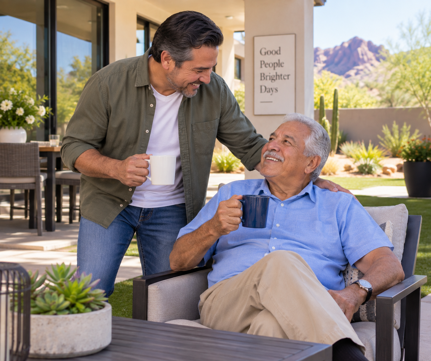 Two men, one sitting outdoors on a patio, smiling and holding a mug, while another man leans over and smiles, also holding a mug, with a scenic mountain view in the background and a sign that says 'Good People Brighter Days' nearby.