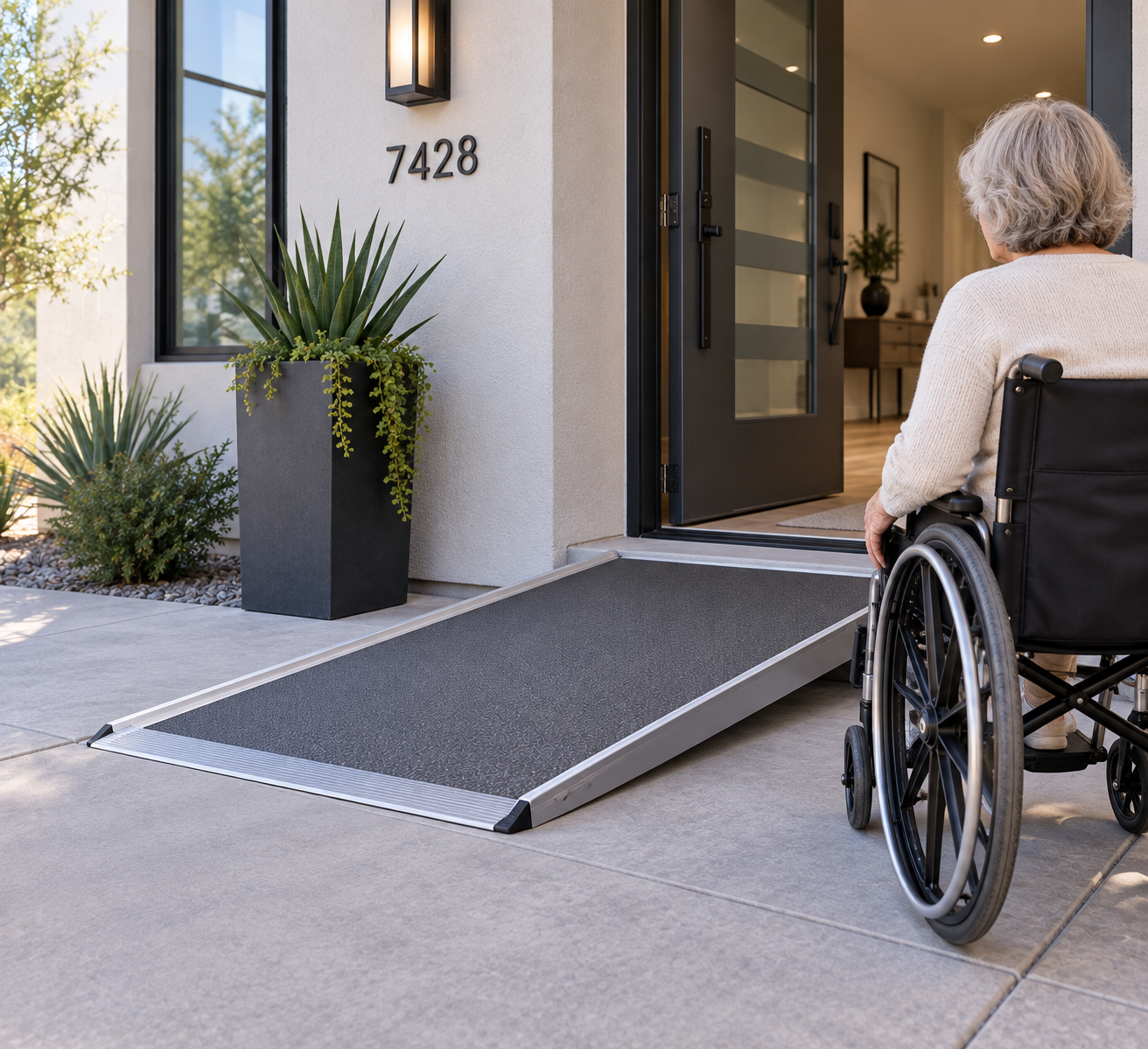 A woman in a wheelchair approaching a house with a ramp at the front door, which has a modern black door and potted plants outside.