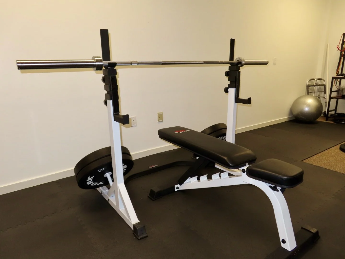 A gym with a York Barbell weight bench, a bar with weight plates, a stability ball, and exercise equipment against a wall.