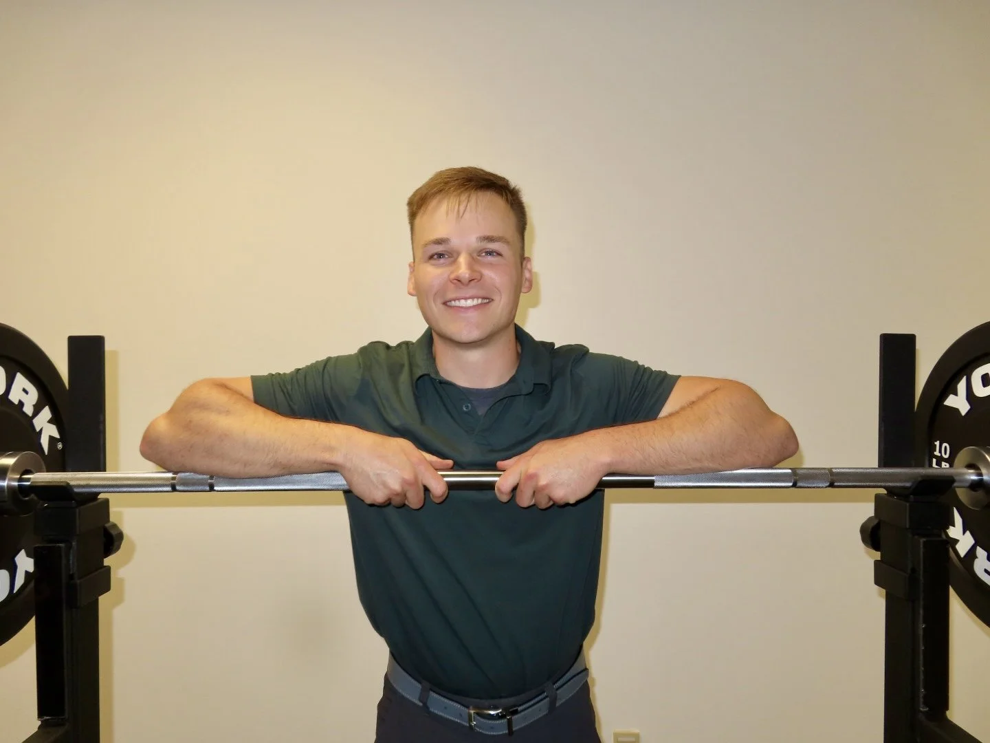 A smiling young physical therapist in a green polo shirt standing behind a barbell on a squat rack, with his arms resting on it, in a gym setting.