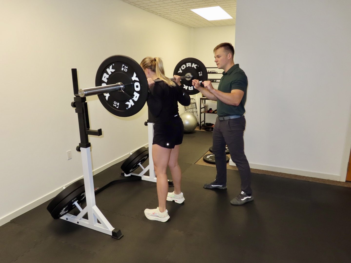 A woman lifting weights with a barbell on her shoulders, supervised by a trainer in a gym