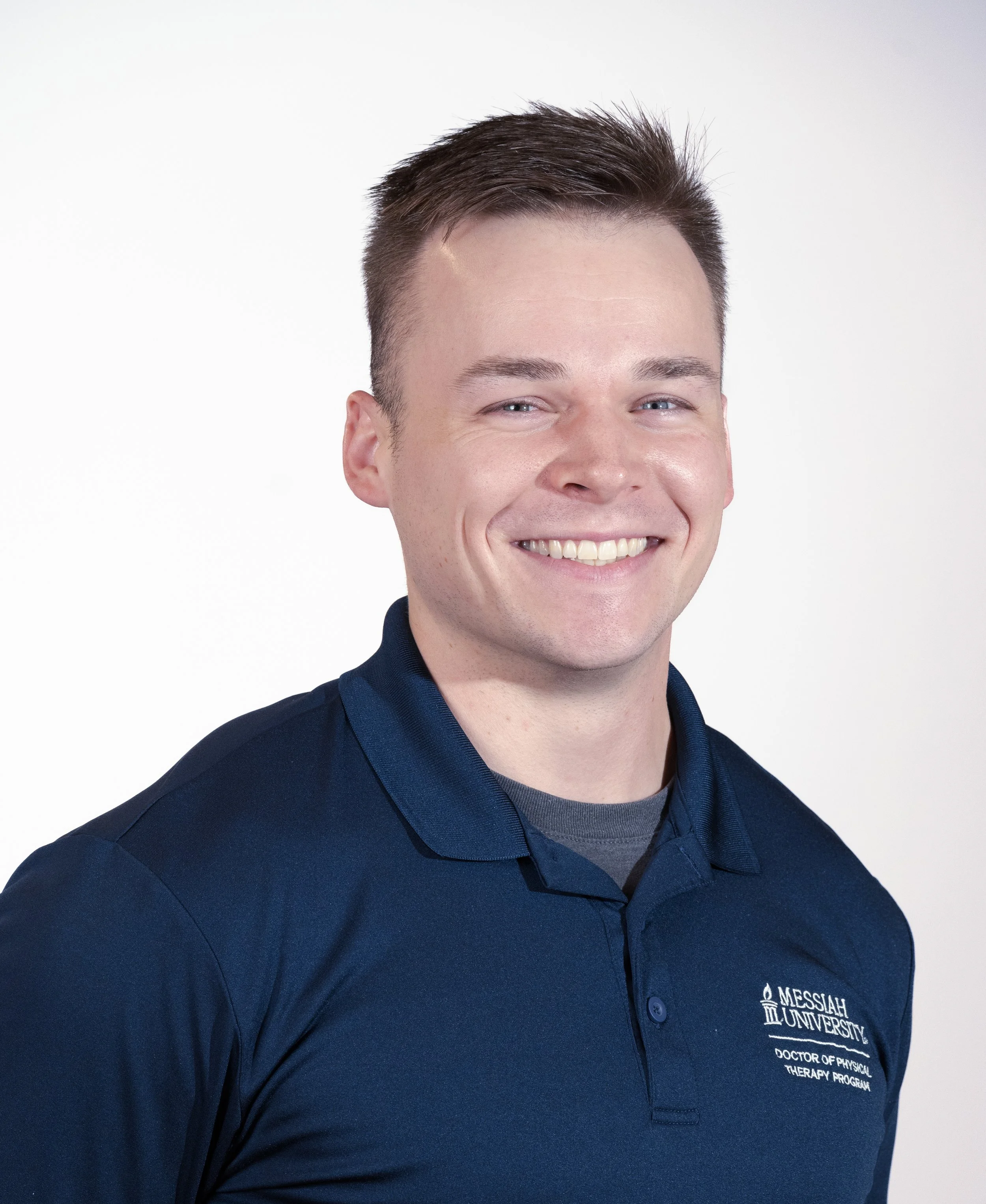 Close-up of a smiling young man wearing a navy blue polo shirt with the logo of Messiah University and the text 'Doctor of Physical Therapy Program' embroidered on it, against a plain white background.