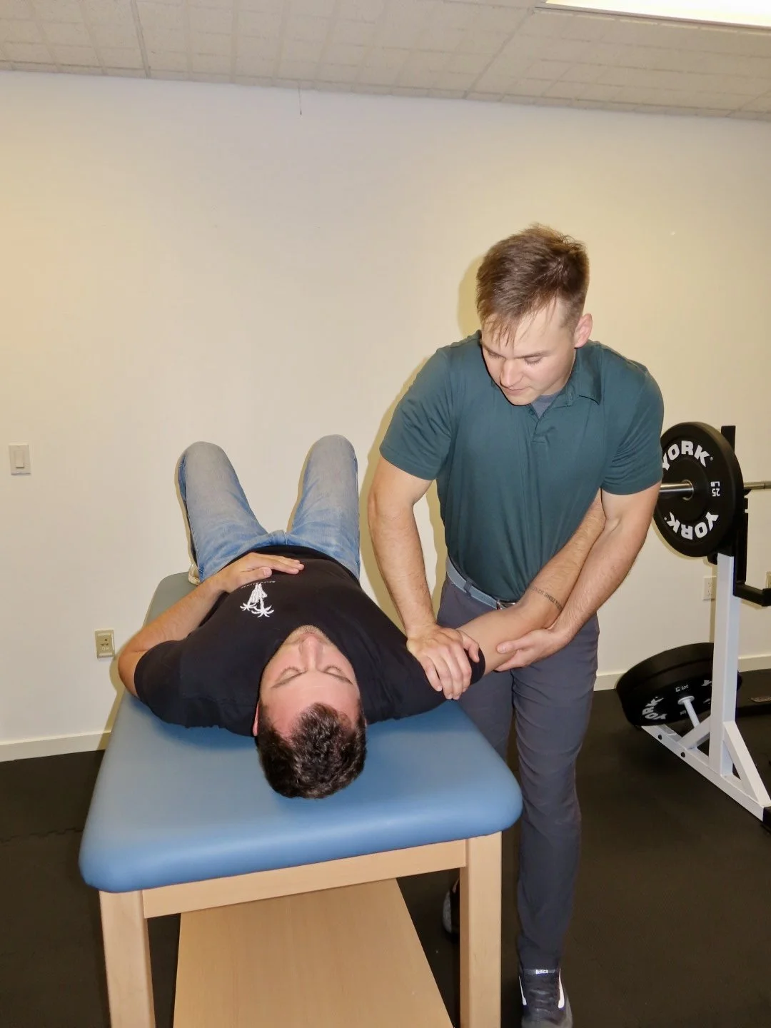 A physical therapist assisting a patient with arm exercises on a therapy table, in a fitness room. The patient lies on their back with their head on the table, holding the therapist's arm, while the therapist stretches their shoulder.