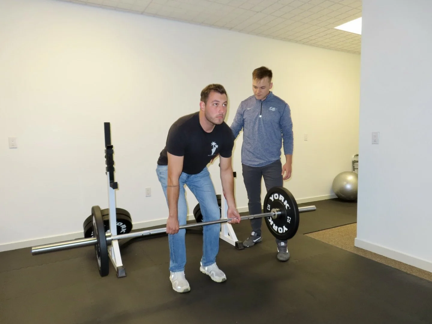 A man in a black t-shirt is practicing deadlifts with a barbell, supervised by a trainer in a blue jacket in a gym with a black floor.