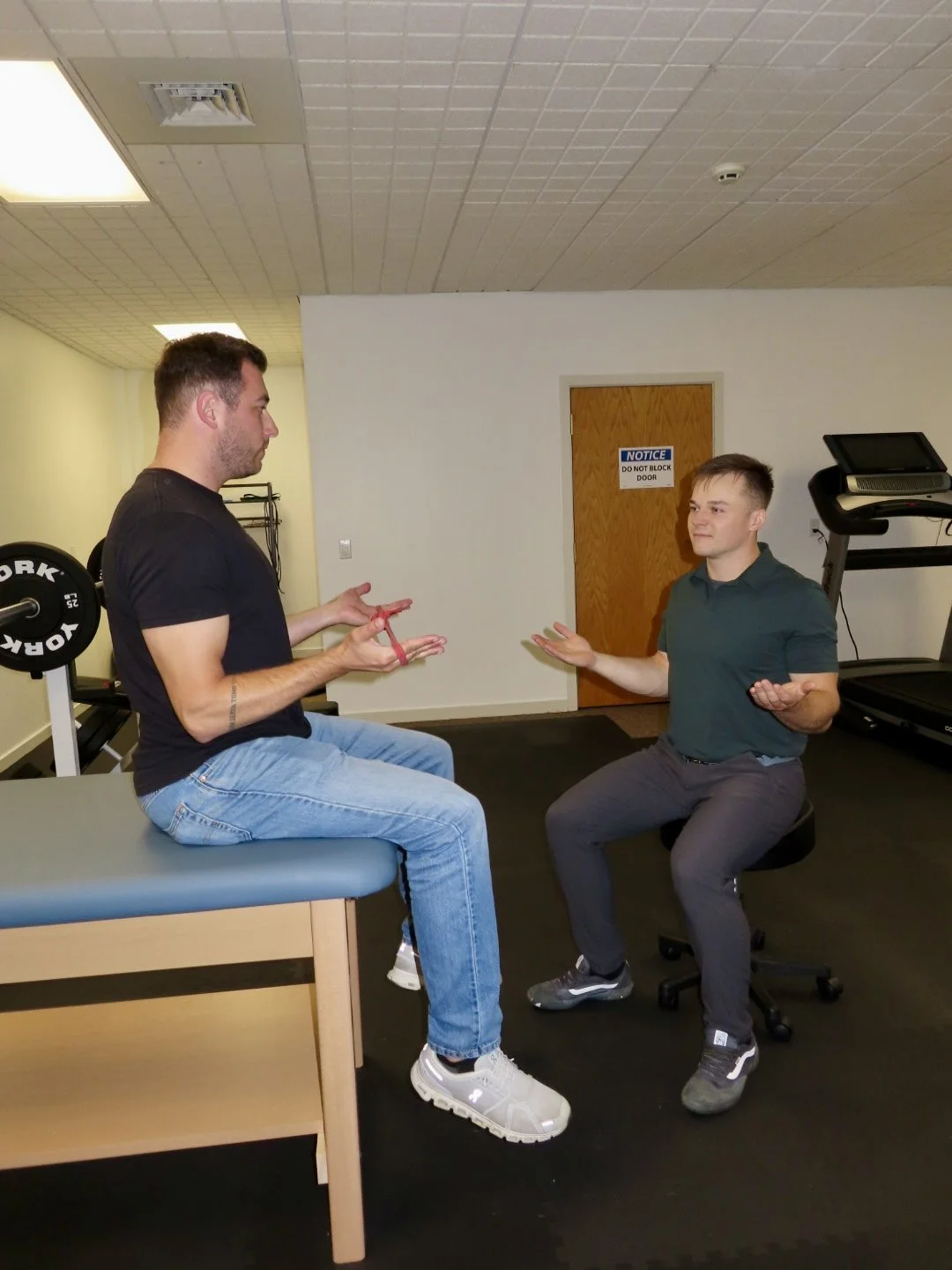 Two men are engaged in a conversation in a gym. One man, sitting on an examination table, holds resistance bands and wears a black t-shirt. The other man, sitting on a stool, gesturing with his hands and wearing a dark green polo shirt and gray pants. The gym equipment, including weights and a treadmill, are visible in the background.