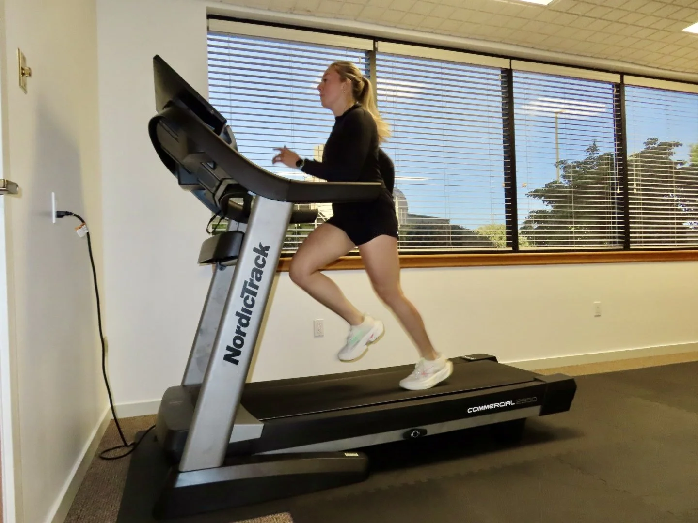 A woman running on a NordicTrack treadmill in a gym or fitness room with large windows.