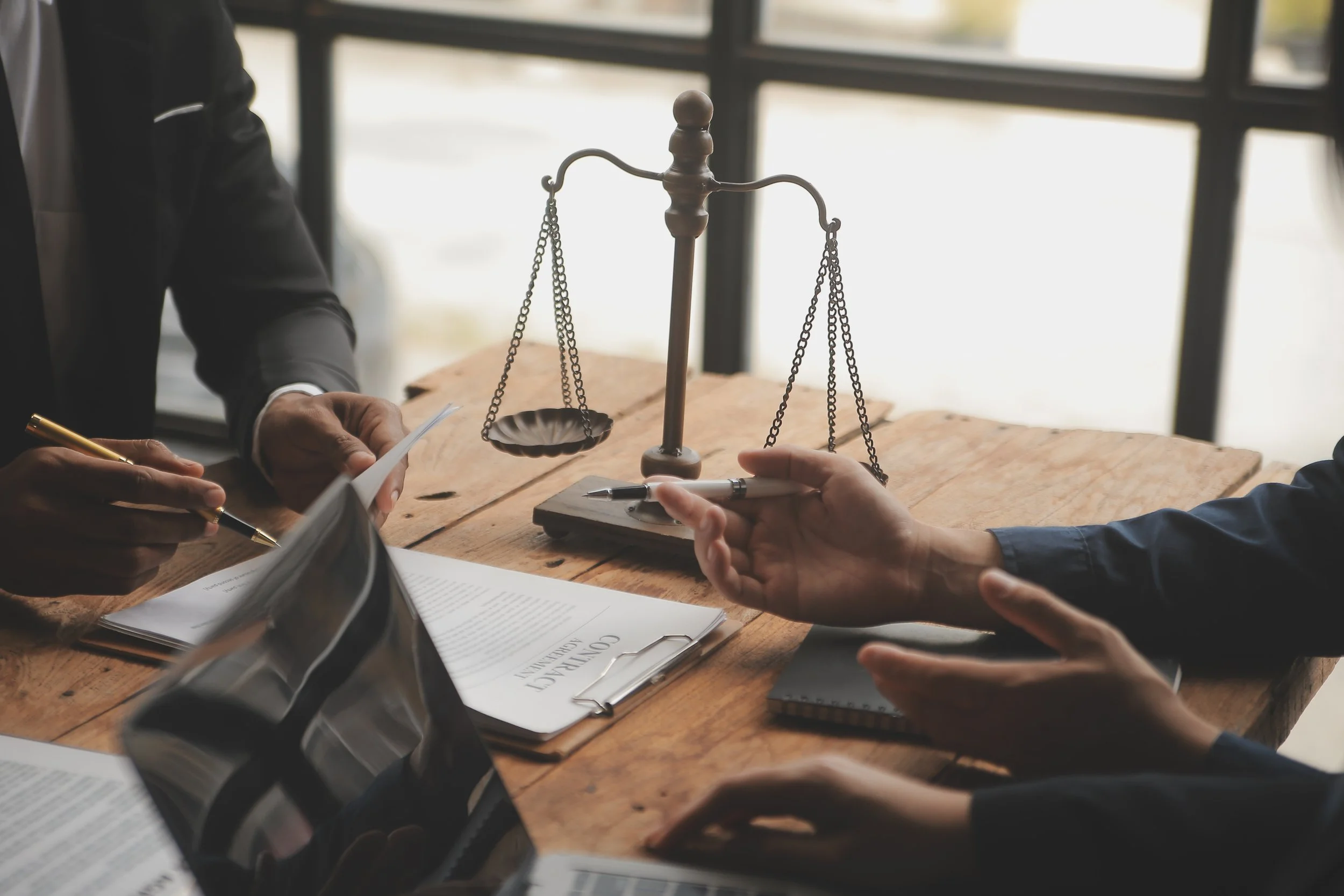 Two people are sitting at a wooden table during a legal negotiation or consultation, with a traditional balance scale on the table between them.