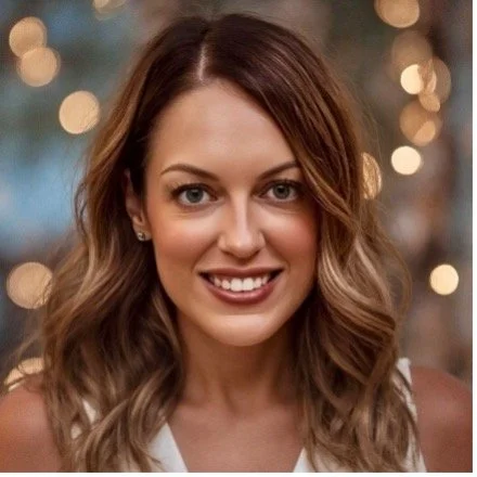 A woman with wavy brown hair and a white top, smiling at the camera with blurred lights in the background.