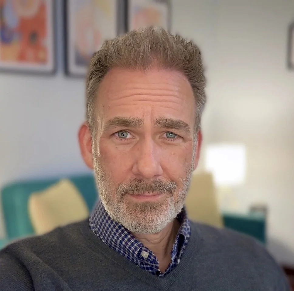 Close-up of a middle-aged man with gray hair and beard, wearing a checkered shirt and dark sweater, in a cozy room with colorful framed pictures and a sofa with yellow cushions in the background.