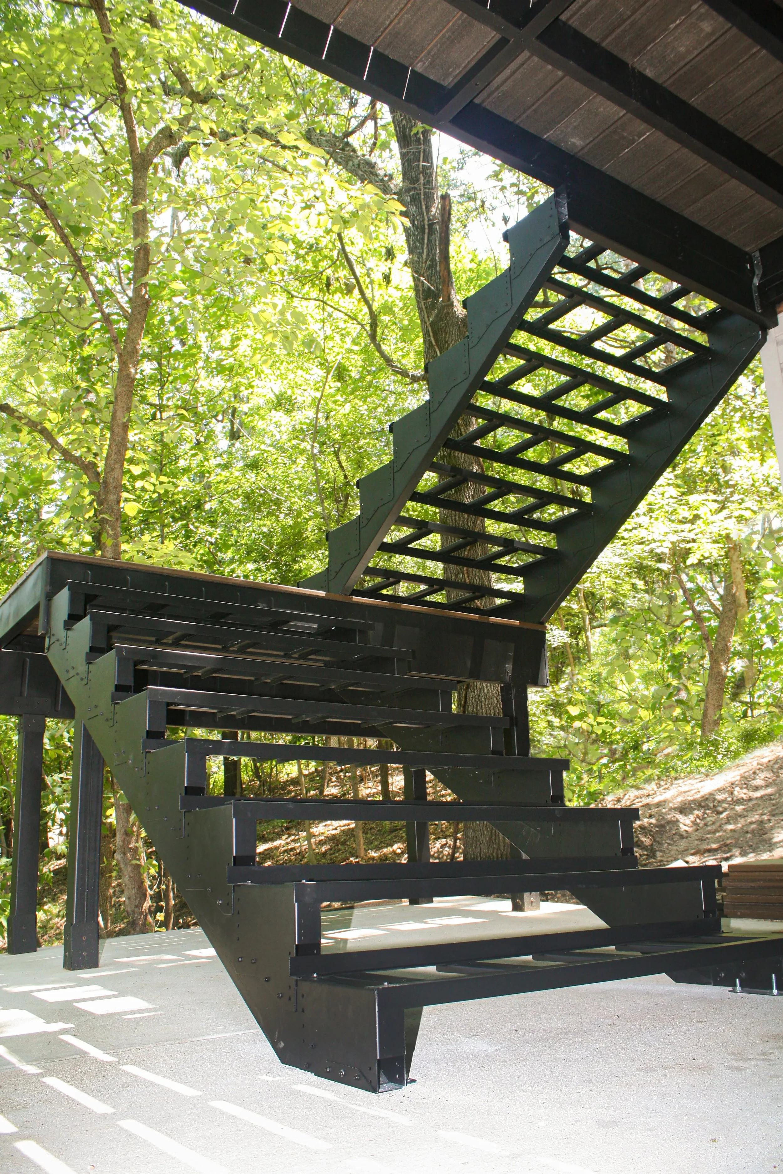 Black metal staircase with open risers and angled design leading up to a building with a dark roof, surrounded by green trees and daylight.