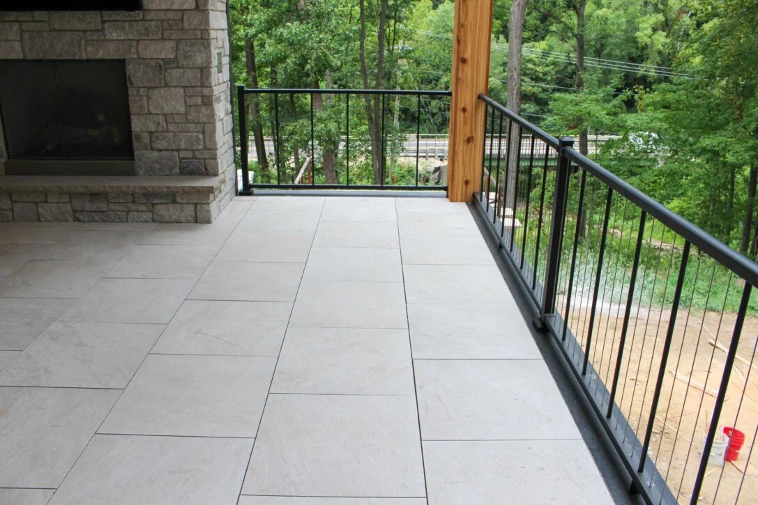 View of a balcony with large white tiles, a black metal railing, a stone fireplace on the left, a wooden post, lush green trees in the background, and a partially visible road.