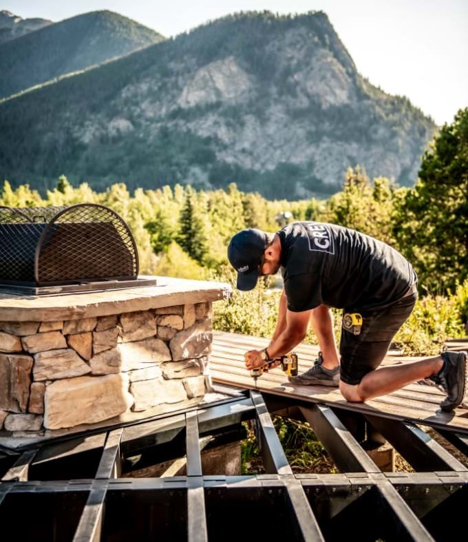 A man working on a wooden deck on a mountain with trees and a large mountain in the background.