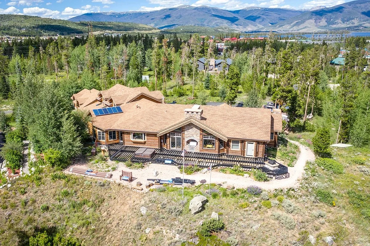A large house with a brown roof, solar panels, and a deck, surrounded by trees, with mountains and a lake in the background.