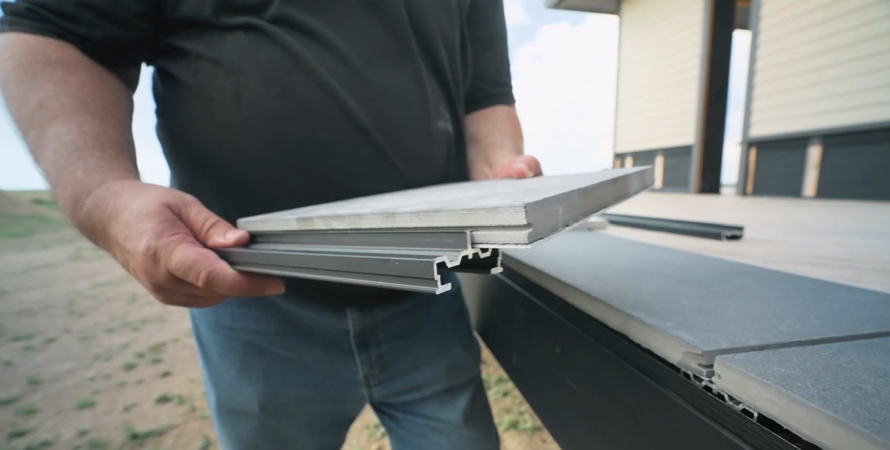 A person holding a piece of gray wood and a gray aluminum siding profile, inspecting or preparing materials outside a building with a clear sky