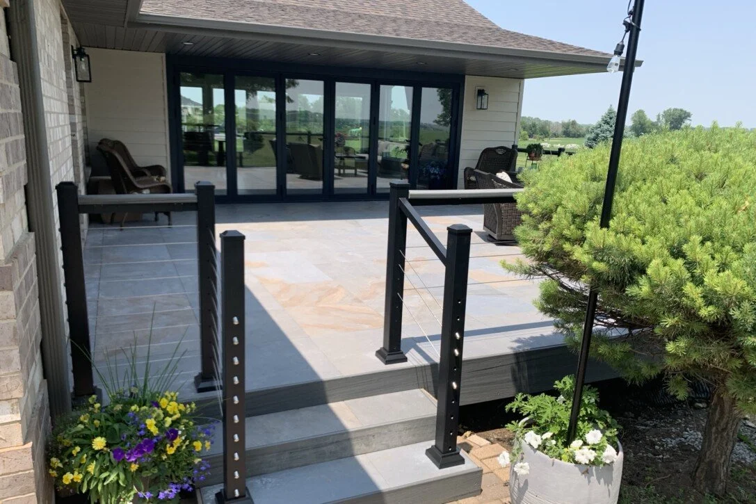 A spacious backyard patio with dark railing, chairs, and a large glass sliding door leading into the house, surrounded by greenery and flower pots.